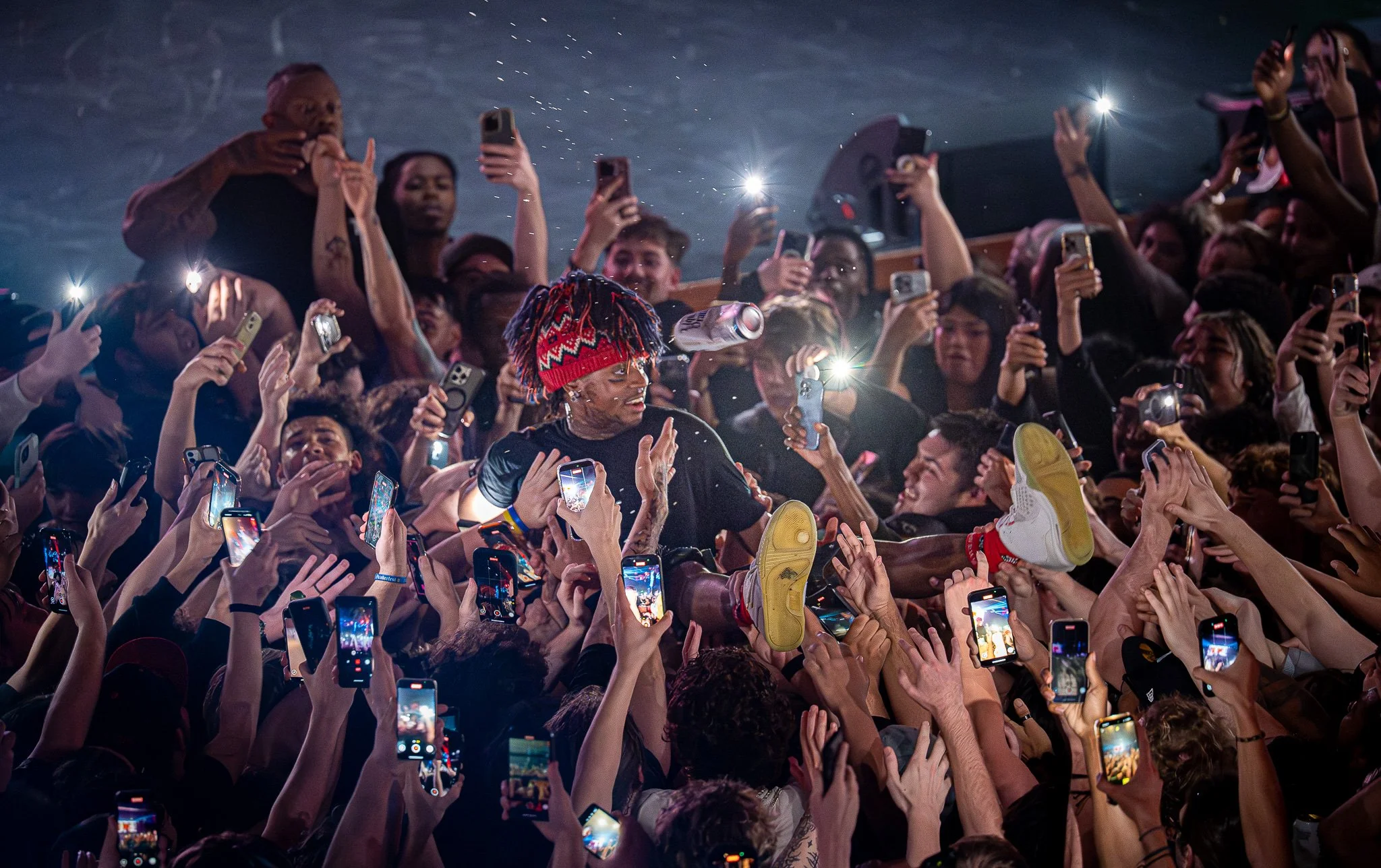 A musician with red and black dreadlocks, wearing a red and black beanie, performing on stage amid a crowd of fans holding up smartphones.