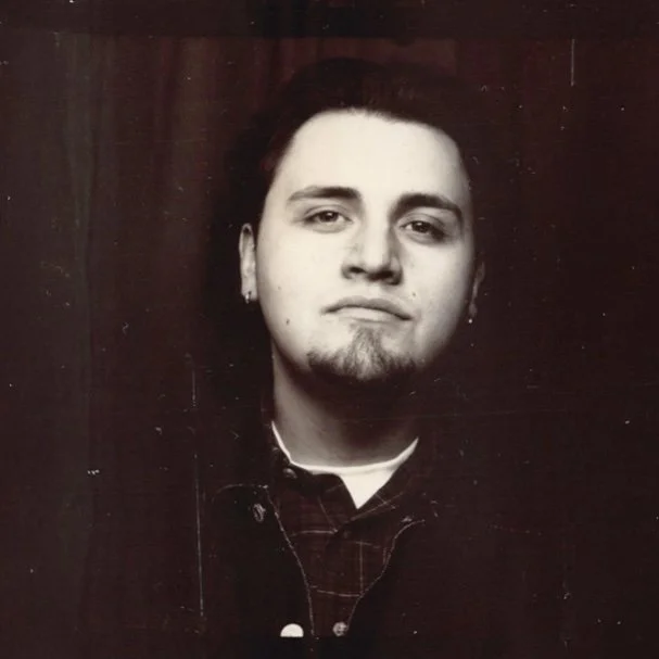 Black and white photo of a young man with short dark hair, slight beard, and piercing, wearing earrings and a checkered shirt.