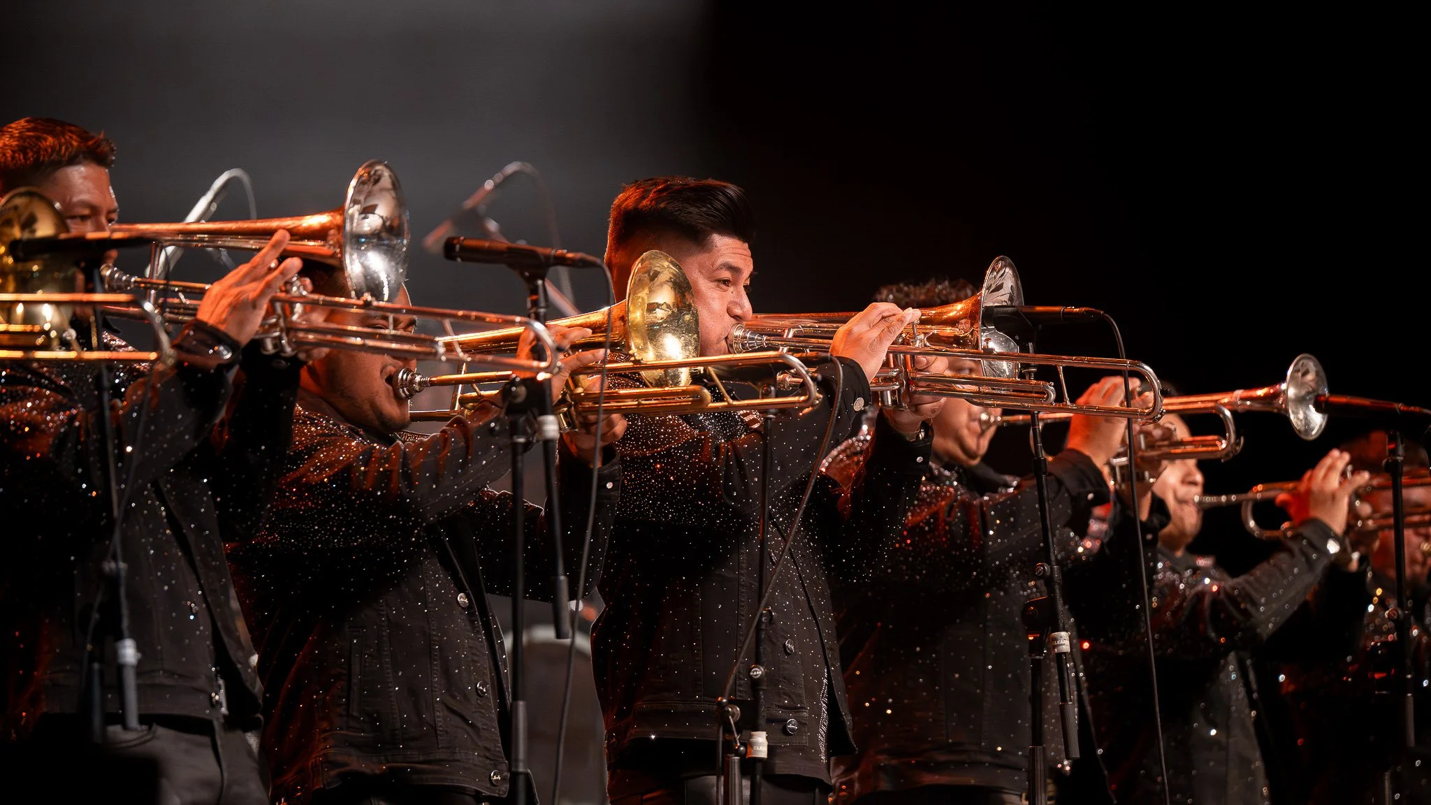 A group of musicians playing trumpets on stage, dressed in black with sparkling details, during a performance.