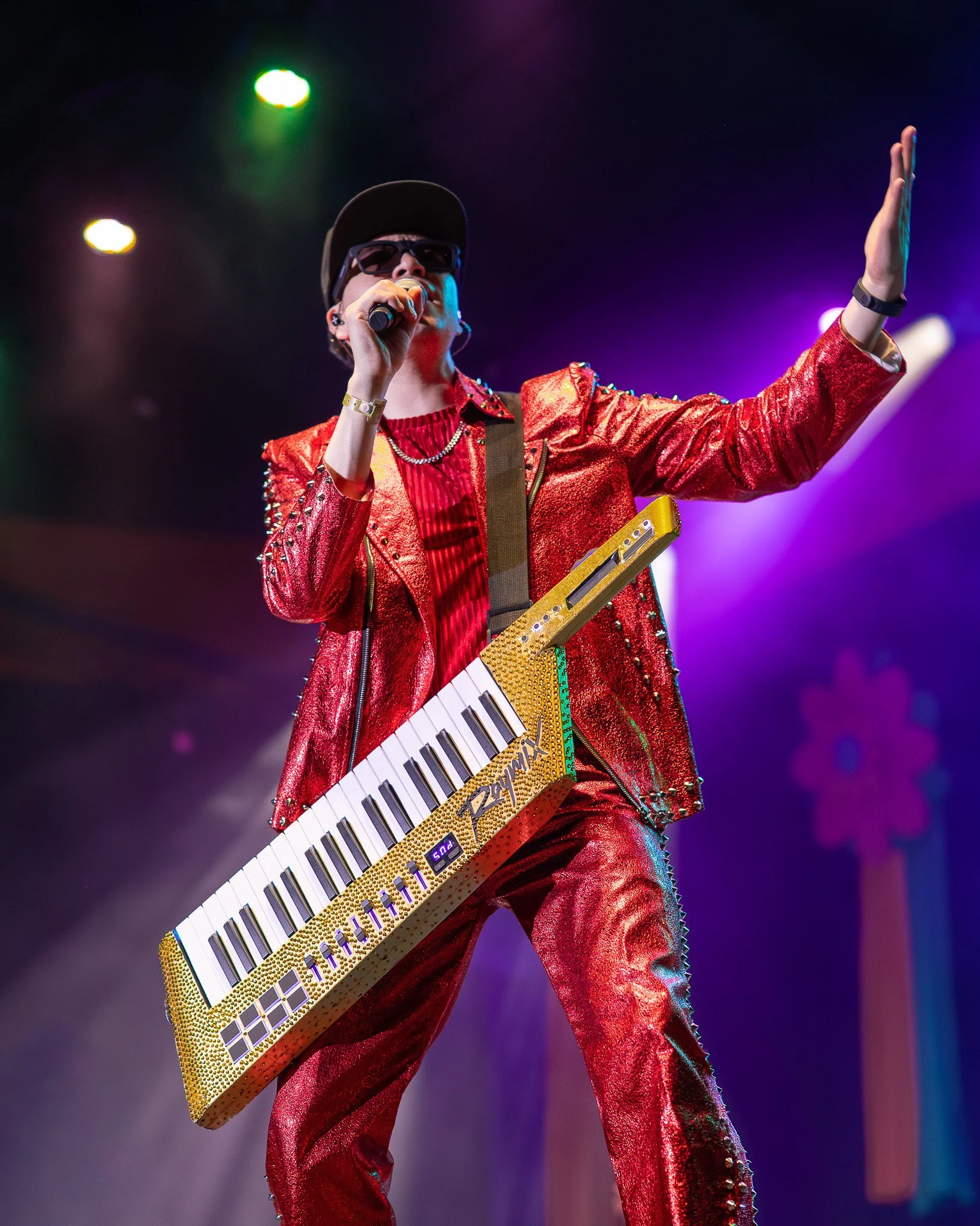 Performer on stage in a red shiny suit, wearing sunglasses and a baseball cap, holding a microphone and a gold-colored keytar keyboard, with colorful stage lights in the background.