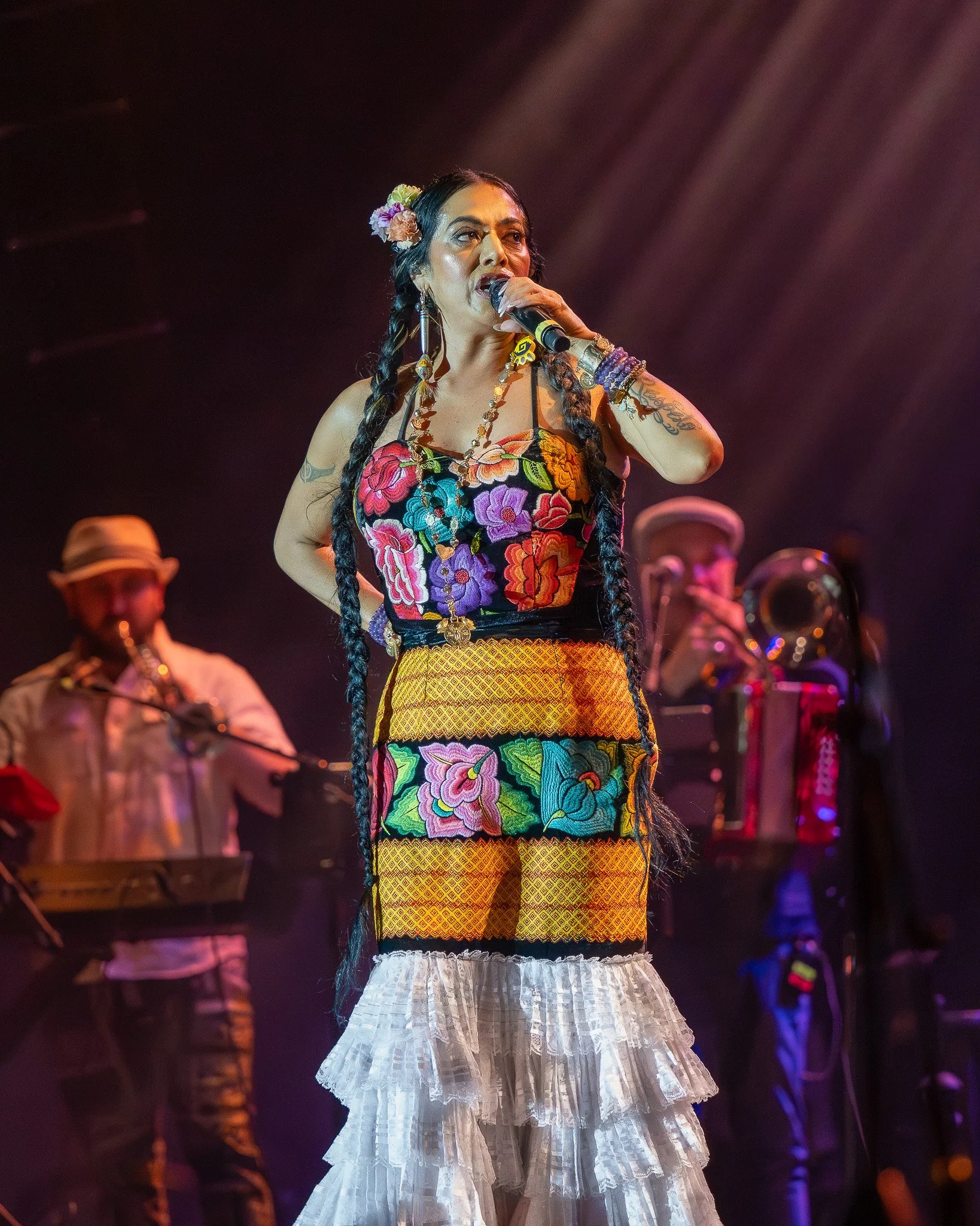 A woman singing into a microphone on stage, dressed in colorful, embroidered Mexican-inspired clothing with braided hair, flanked by musicians playing instruments in the background.
