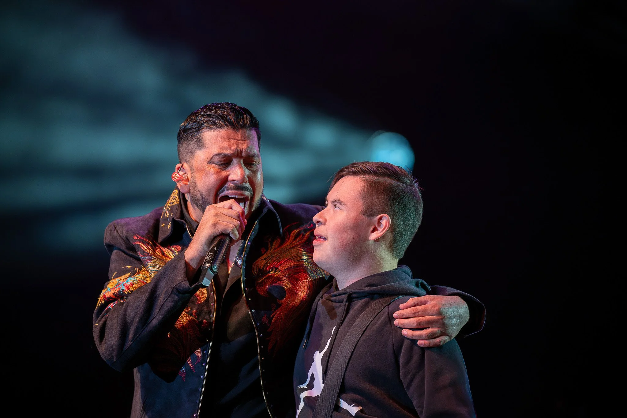 A man singing into a microphone with his eyes closed, having an emotional expression, being comforted by a boy who looks up at him, with a dark background and lighting from one side.