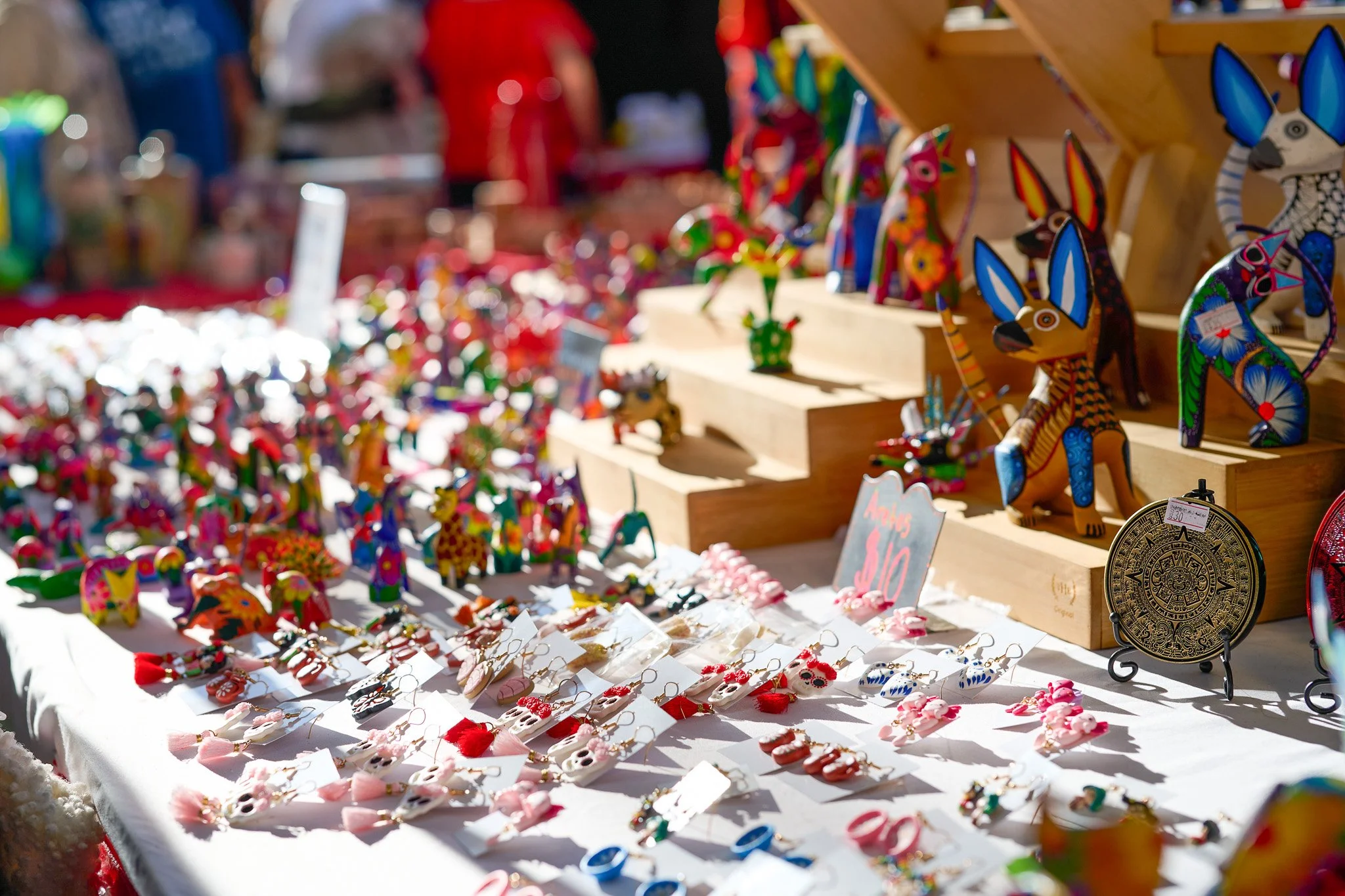Colorful handcrafted figurines and jewelry on a market stall, with wooden animal figures and small decorative items displayed on a white tablecloth and wooden shelves.