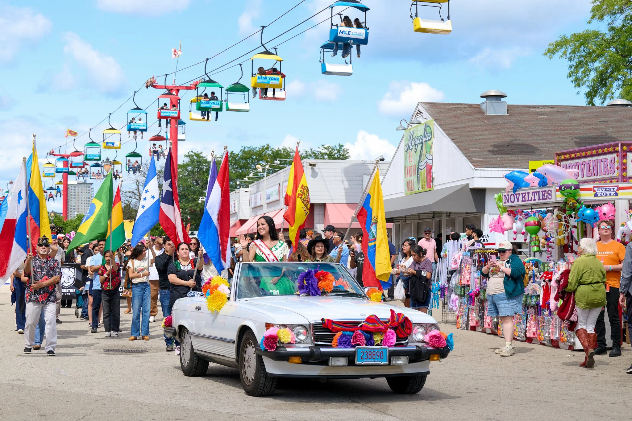 A parade with a float decorated with flowers and costumed women waving flags, followed by a crowd of spectators. The scene is set at a fairground with souvenir stalls and a skyride in the background.