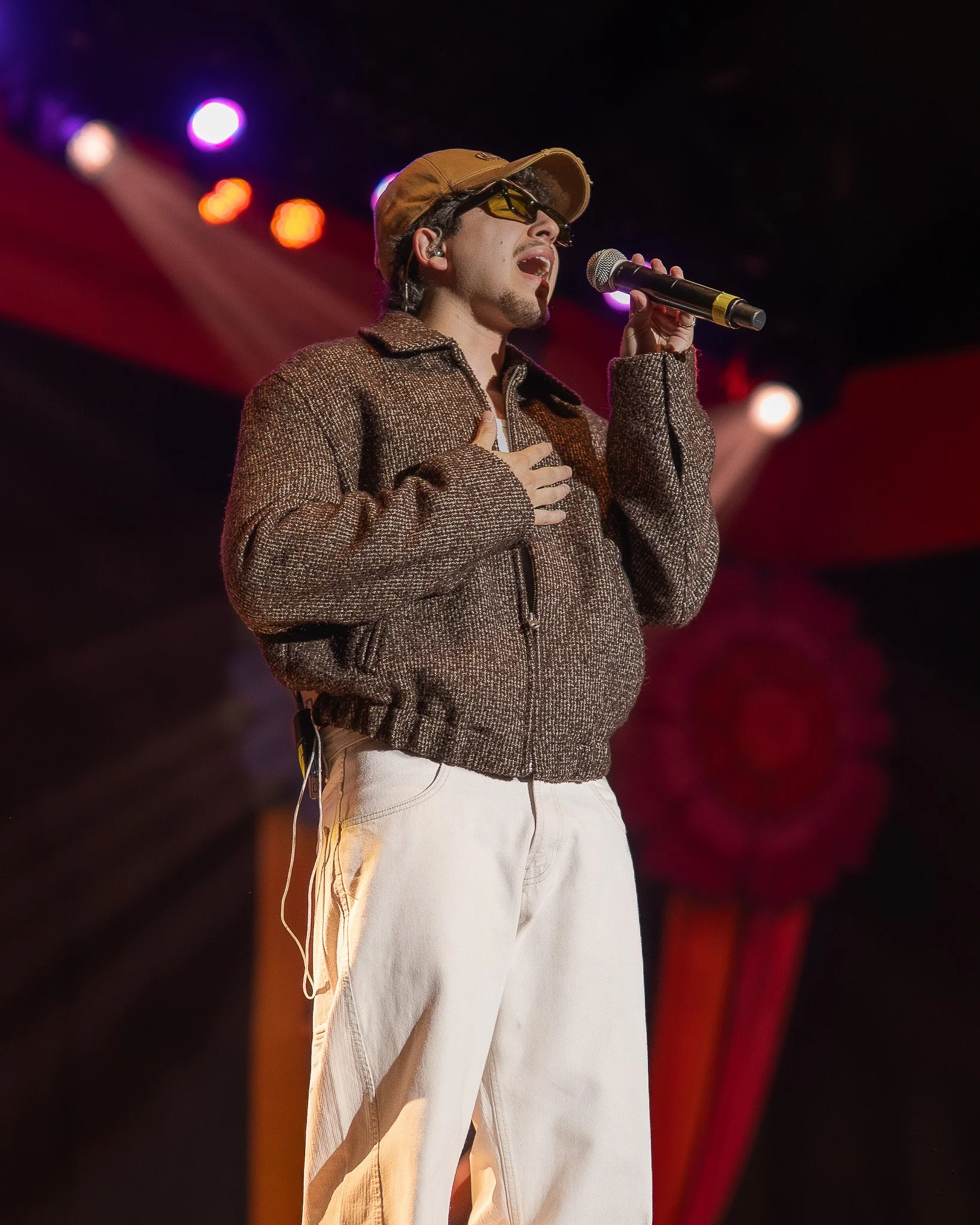 Young man singing on stage with hand on chest, wearing sunglasses, brown cap, brown jacket, and cream-colored pants.