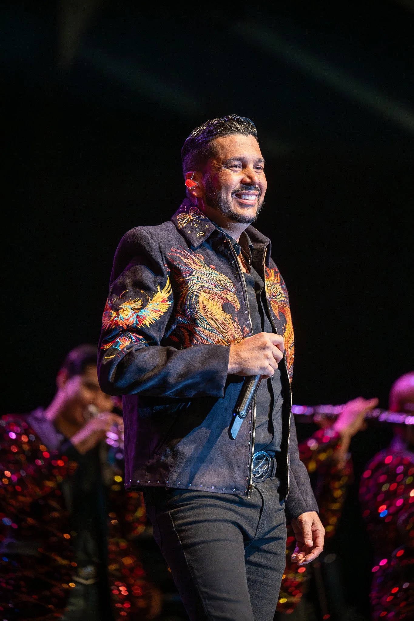 Performer on stage holding microphone, wearing black jacket with colorful embroidered dragons and phoenixes, smiling amid backup musicians in shimmering outfits.