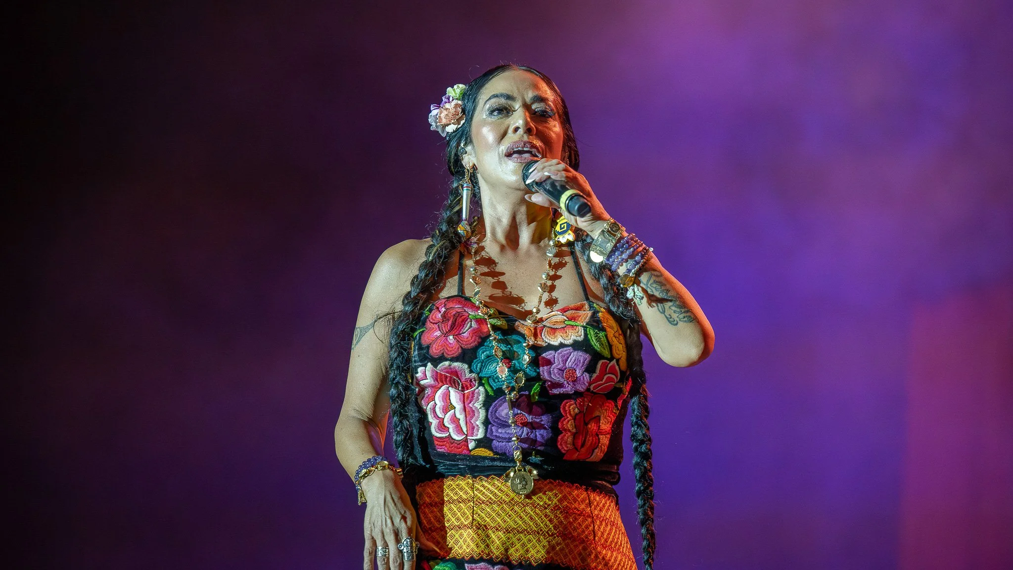 A woman performing on stage, holding a microphone, wearing a colorful embroidered top, jewelry, and flowers in her hair, with purple stage lighting in the background.