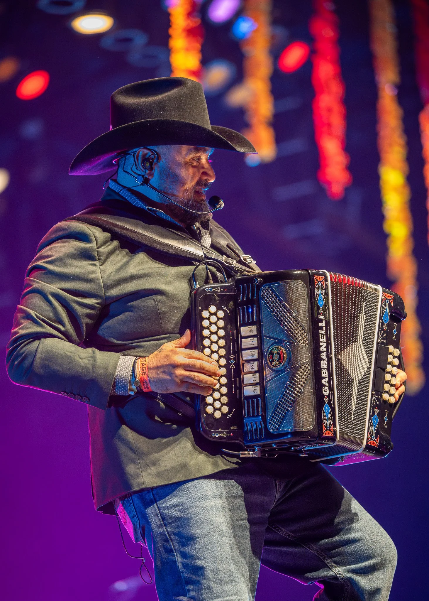 A man wearing a cowboy hat and a jacket, playing an accordion on stage with colorful lights in the background.
