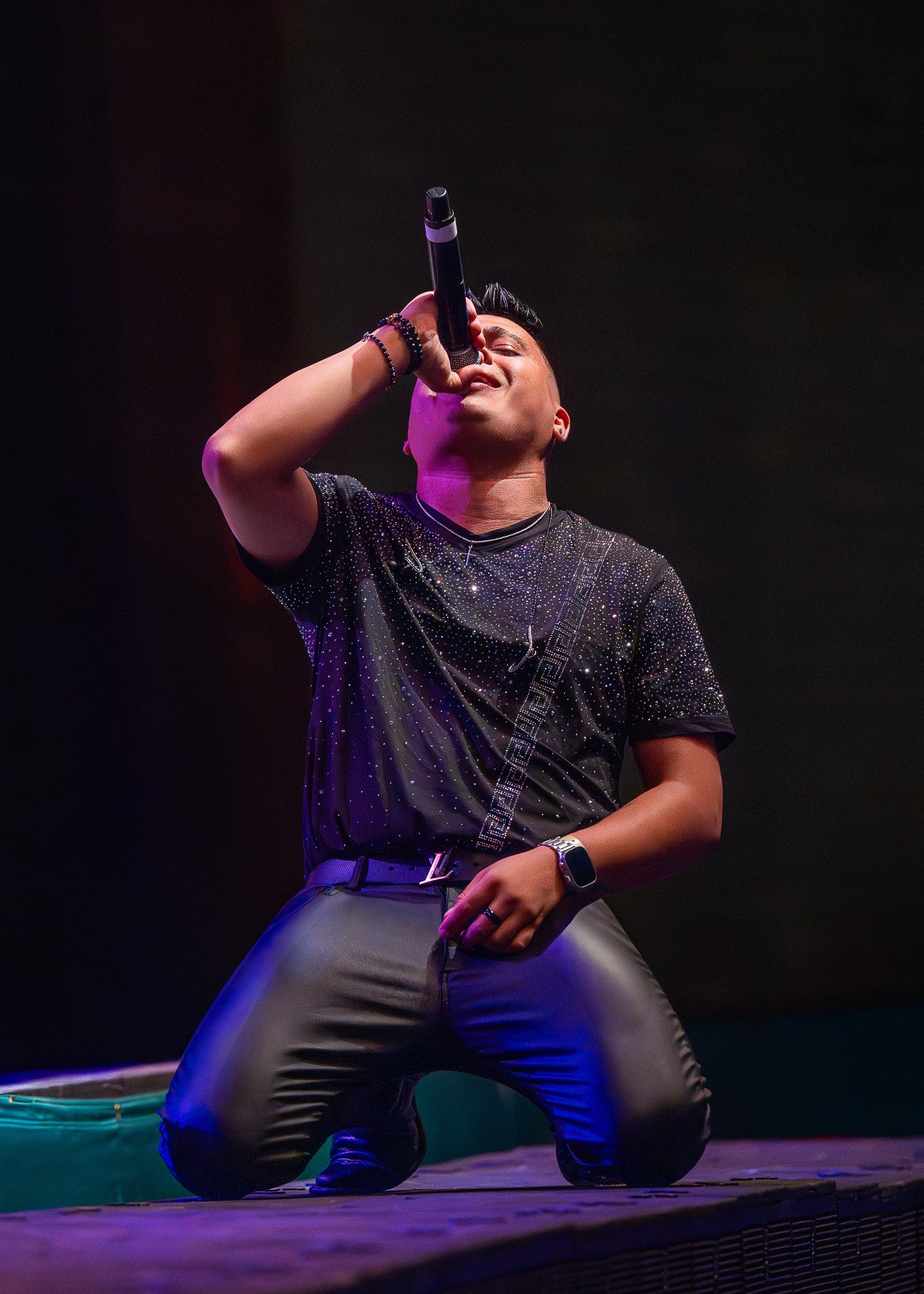 A male singer kneeling on stage, holding a microphone to his mouth, wearing a black shirt with glitter and leather pants, performing under stage lights.