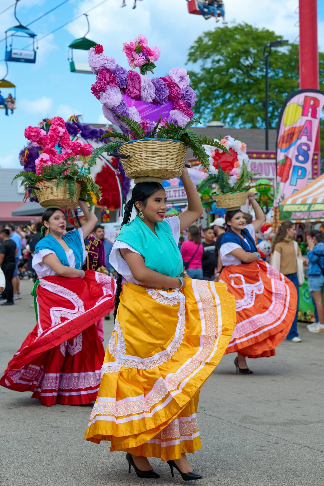 Women in colorful traditional dresses participating in a cultural parade, carrying baskets of flowers on their heads, with carnival rides and other festival attractions in the background.
