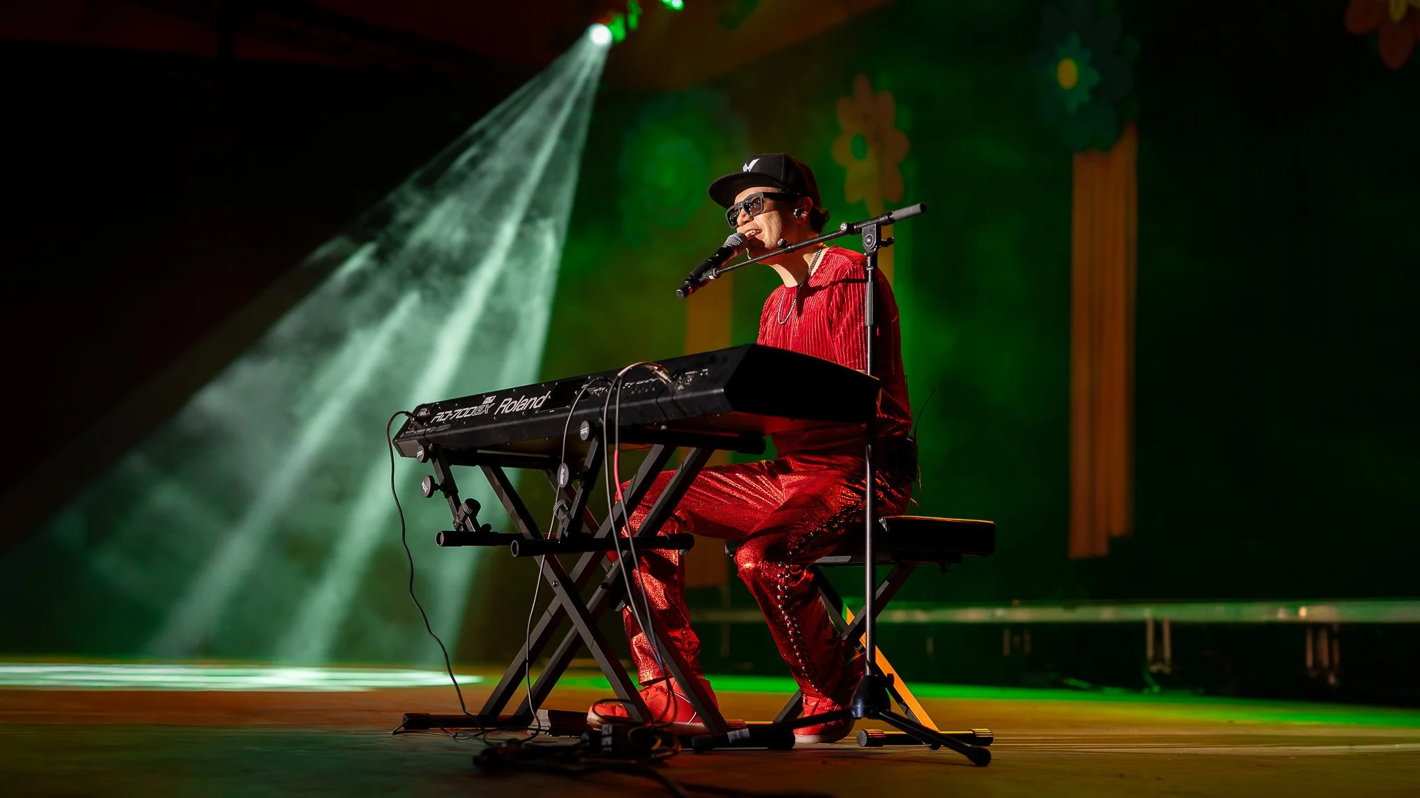 Performer in red outfit playing a keyboard vocals on stage with green and yellow backdrop, wearing sunglasses and black cap.