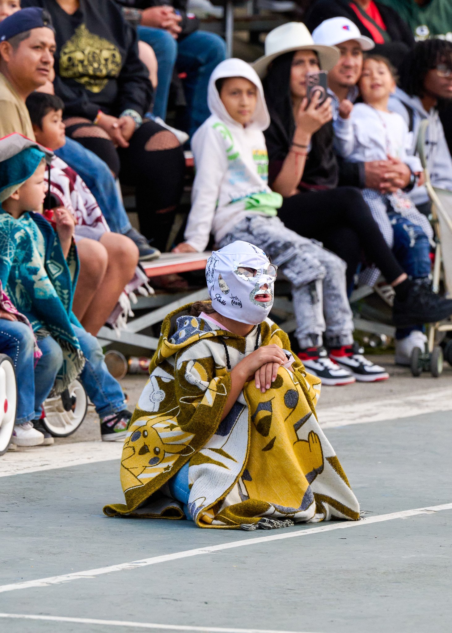 A person in a Pikachu blanket with a white mask sits on the ground in front of an audience, at an outdoor event.