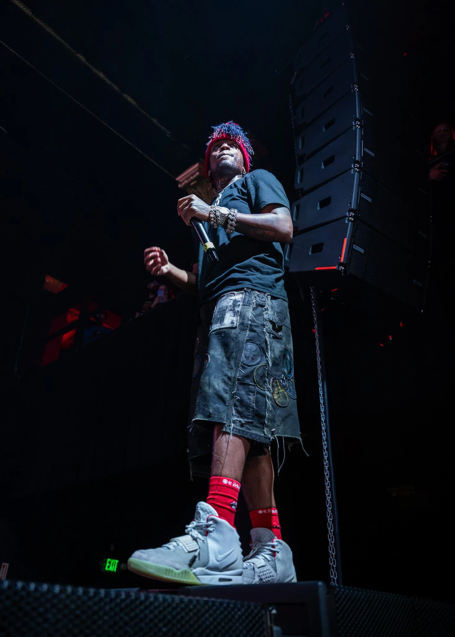 A male performer stands on stage holding a microphone, wearing a black t-shirt, distressed patchwork shorts, red crew socks, and white sneakers, with red hair and accessories including bracelets, in a dark concert setting.