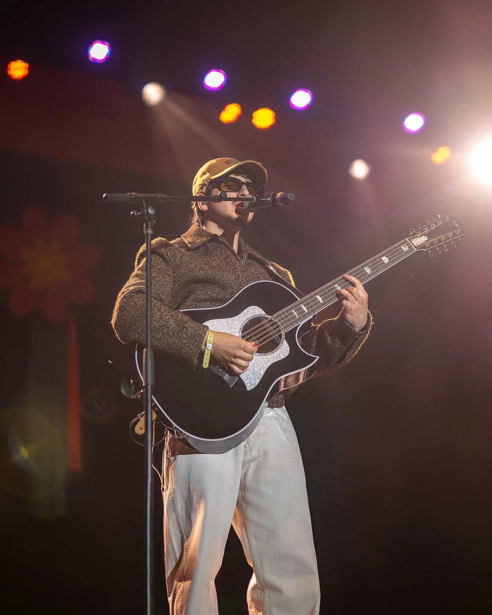 A man performs on stage with a guitar, wearing sunglasses, a cap, and a brown jacket, illuminated by stage lights.