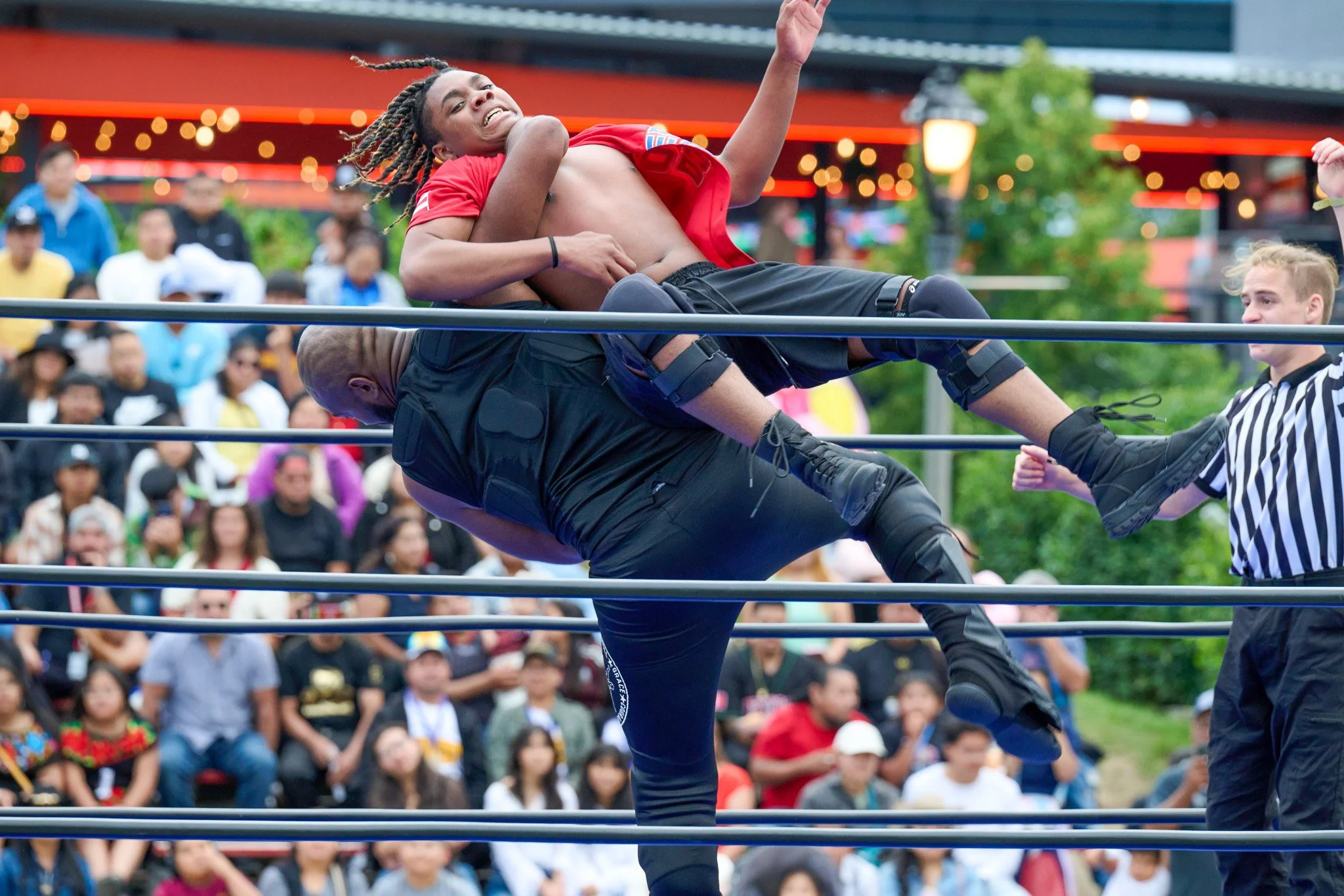 A professional wrestling match with two wrestlers in the ring, one lifting the other, while a referee watches, and an audience seated outdoors in the background.