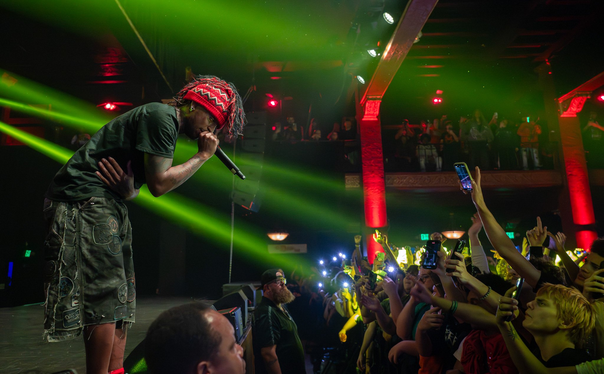 A male performer with dreadlocks, wearing a red knit hat, black T-shirt, and camouflage shorts, sings into a microphone on stage during a concert. The audience in front of him is cheering and taking photos with their smartphones. The venue is decorated with red and green lighting and ornate architectural elements.