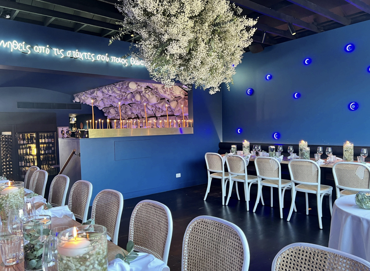 A restaurant interior with blue walls decorated with blue lights and a neon sign in Greek. There are wooden tables set with white napkins, glasses, and cutlery, surrounded by white chairs with woven cane backs. The ceiling has exposed wooden beams, and a projector is mounted on it.