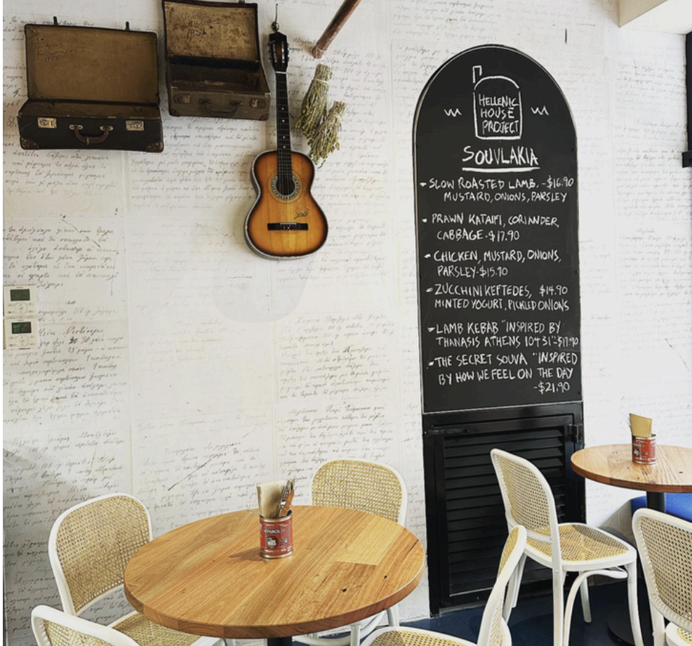 Storefront with a sign that reads 'The Hellenic House Project', featuring outdoor seating with chairs and tables, and signs for property sales and leasing by David Marshall Property.