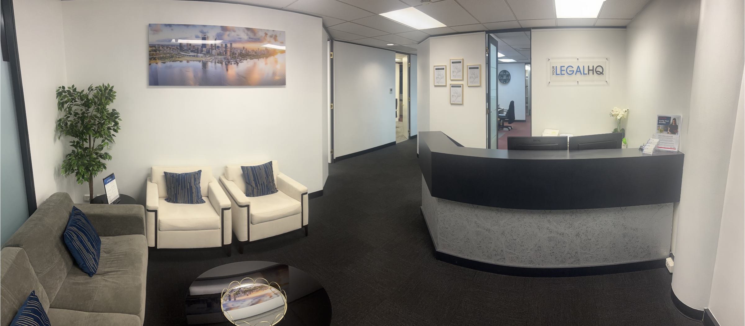 An office reception area with a curved black desk, a sign reading 'Your LegalHQ,' a seating area with gray and white chairs, a coffee table, a potted plant, and framed pictures on the wall.