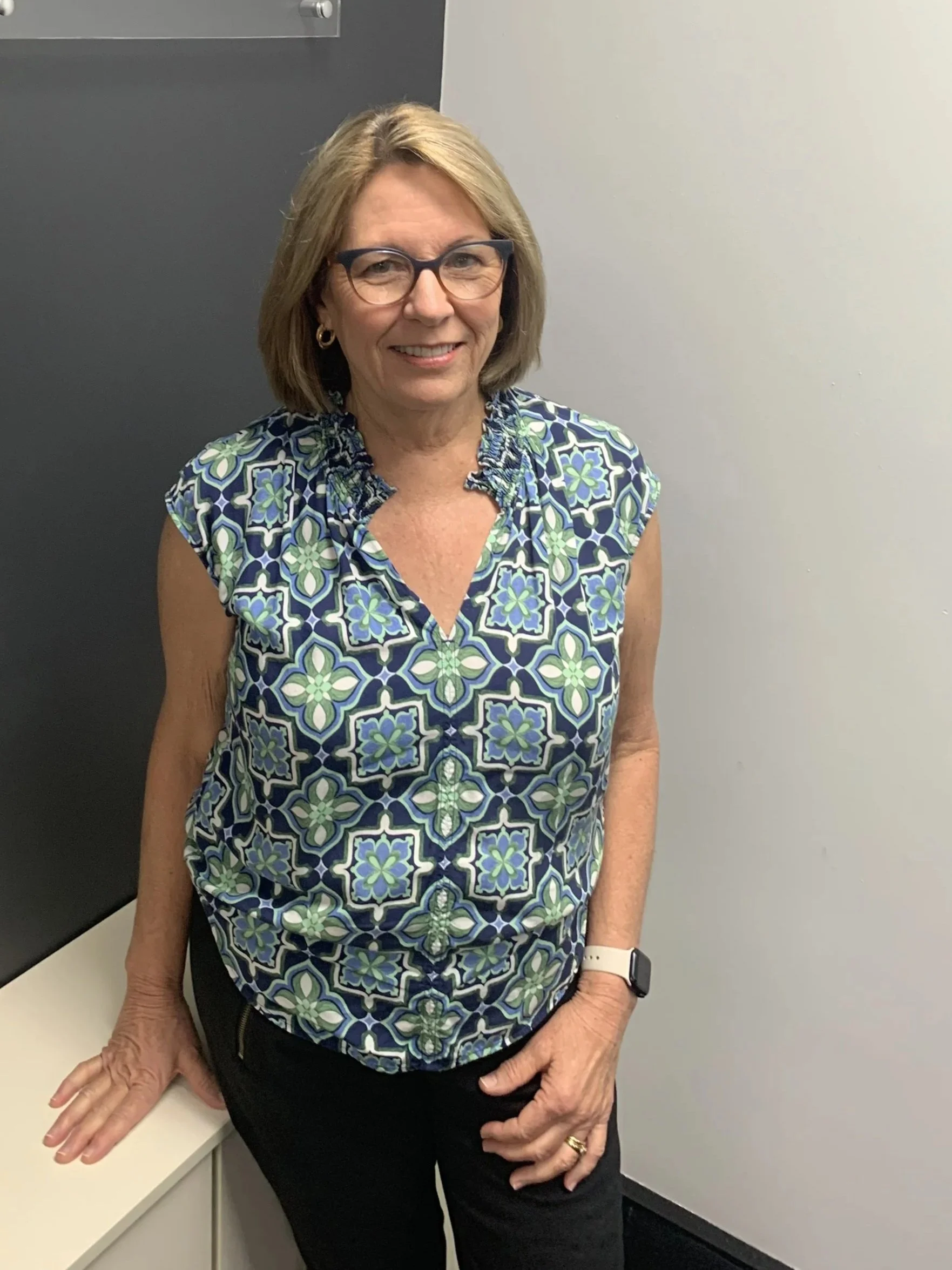 A woman with glasses, blonde hair, wearing a patterned sleeveless blouse, standing in front of a sign that reads 'LEGALHQ' in an office or reception area.