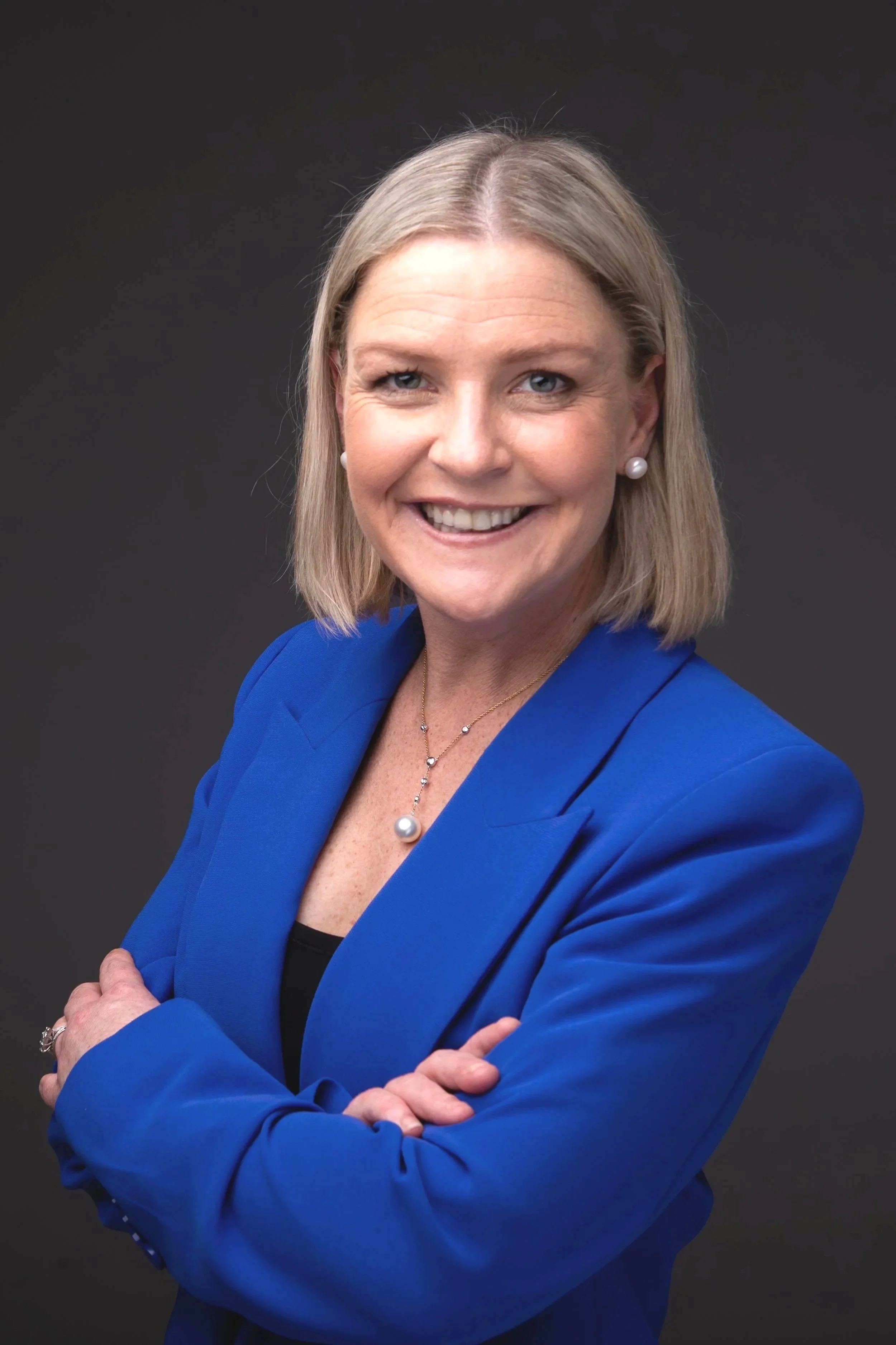 A smiling woman with blonde hair in a bob cut, wearing a blue blazer, pearl earrings, a pearl necklace, and rings, standing against a dark background.