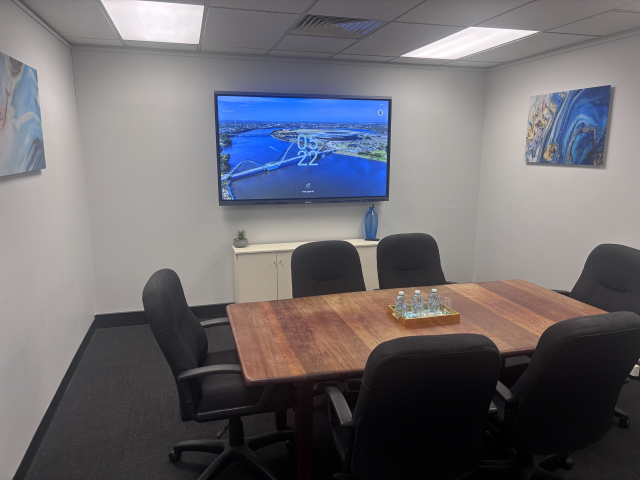 Conference room with a wooden table, six black chairs, a wall-mounted screen displaying a cityscape and water, and abstract art on the walls.