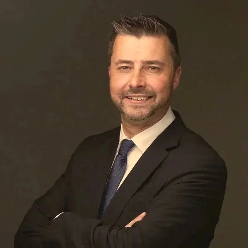 Headshot of a smiling man with short, dark hair and a beard, wearing a black suit, white shirt, and blue tie against a dark background.