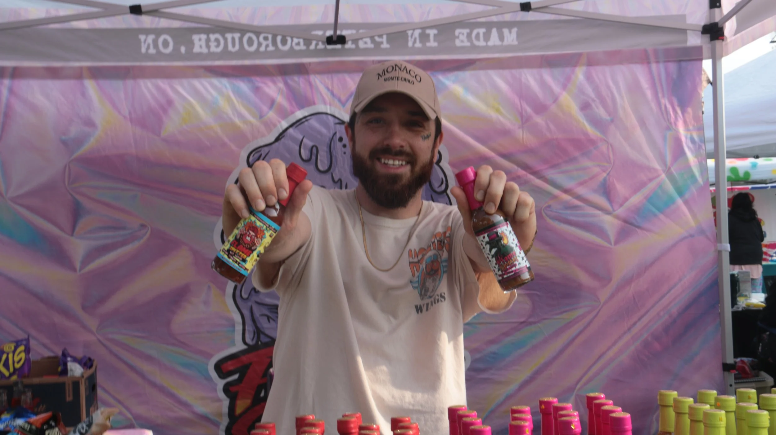 A smiling man with a beard and tattoos on his face holding two bottles of hot sauce, standing in front of a colorful backdrop with a large cartoon face. There are rows of bottles with pink caps and yellow caps on a table in front of him, at an outdoo