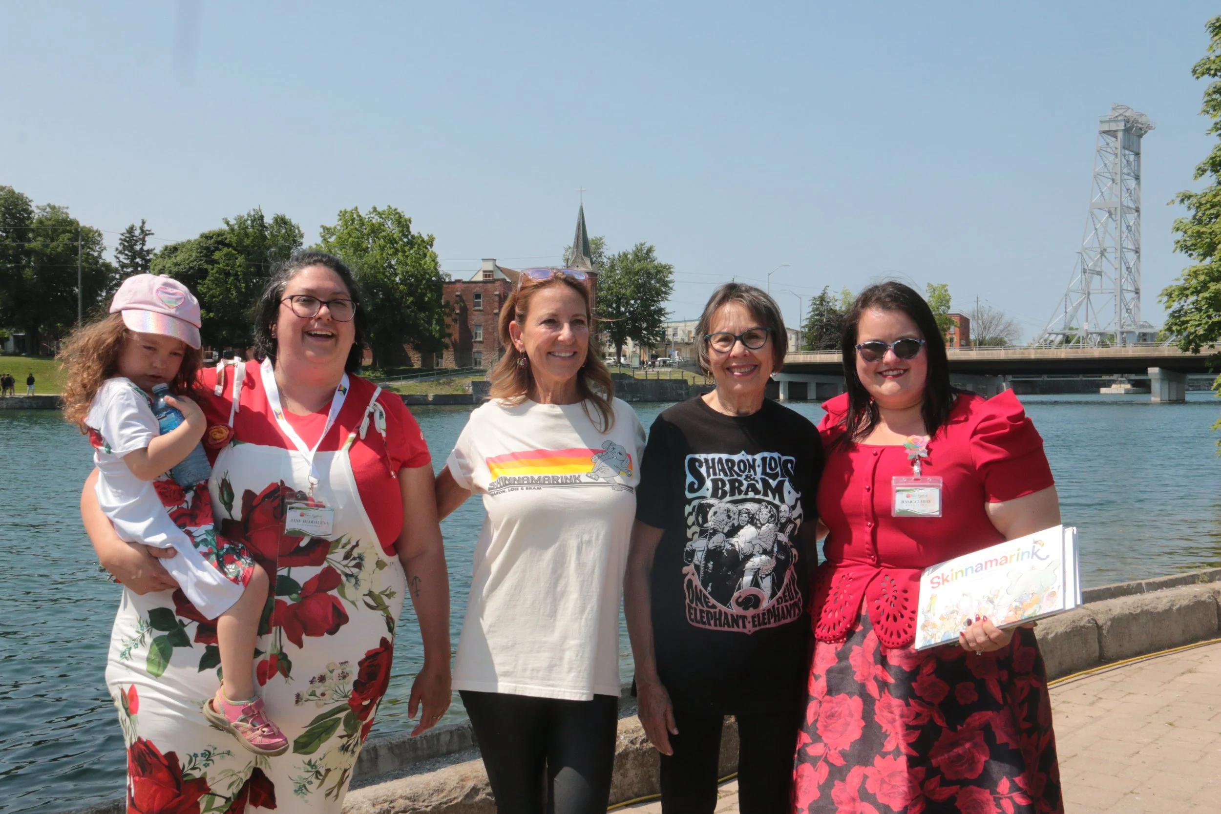 Group of five women standing by a river, smiling for a photo, with one woman holding a young girl, during daytime with blue sky and trees in the background.