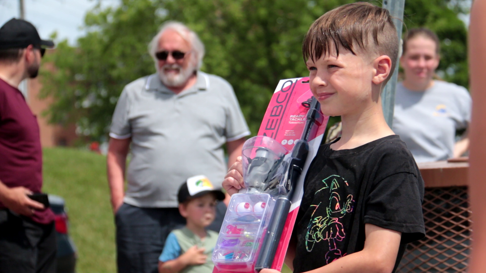 A young boy holding a fishing rod and tackle box, smiling outdoors at a gathering with adults and children, with trees in the background.