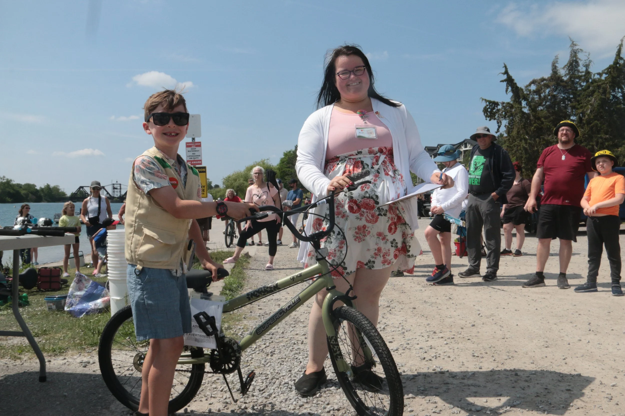 A young boy with sunglasses and a beige vest holding a bicycle, standing next to a woman in a floral dress and white blazer, at an outdoor event by a body of water, with several people in the background.