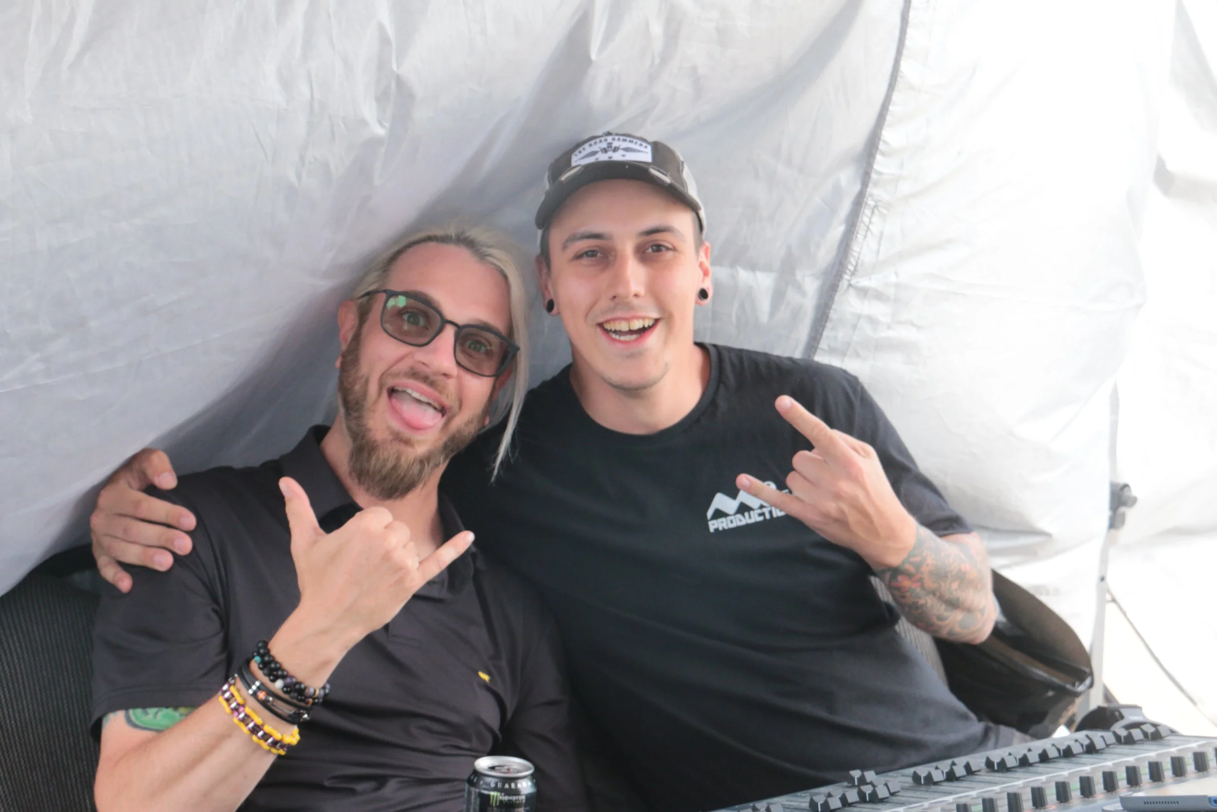 Two men sitting close together smiling and making hand gestures, one wearing glasses and the other with tattoos on his arm, in a white tent or canopy with electronic equipment in front of them.