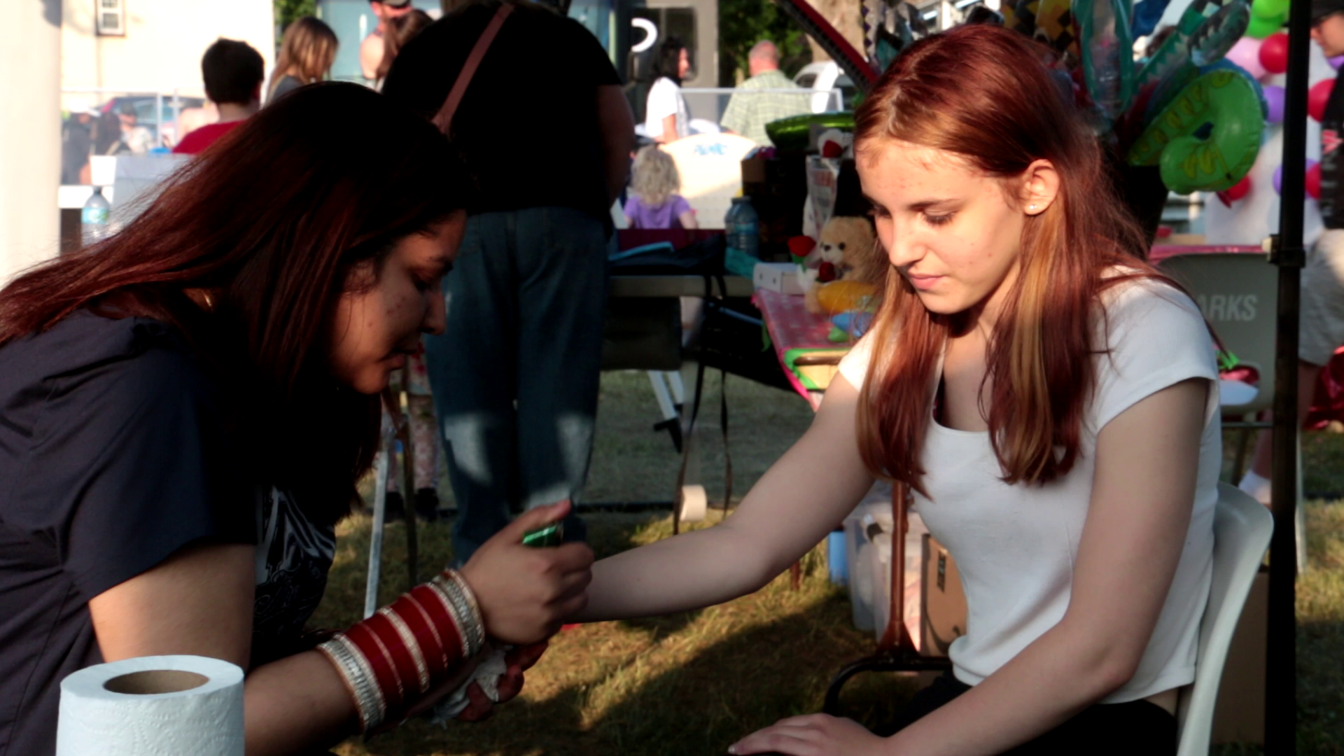 Two young women at an outdoor event, one sitting with her arm extended and the other drawing on it with a marker, smiling and engaging in a face painting activity, with a booth and other people in the background.