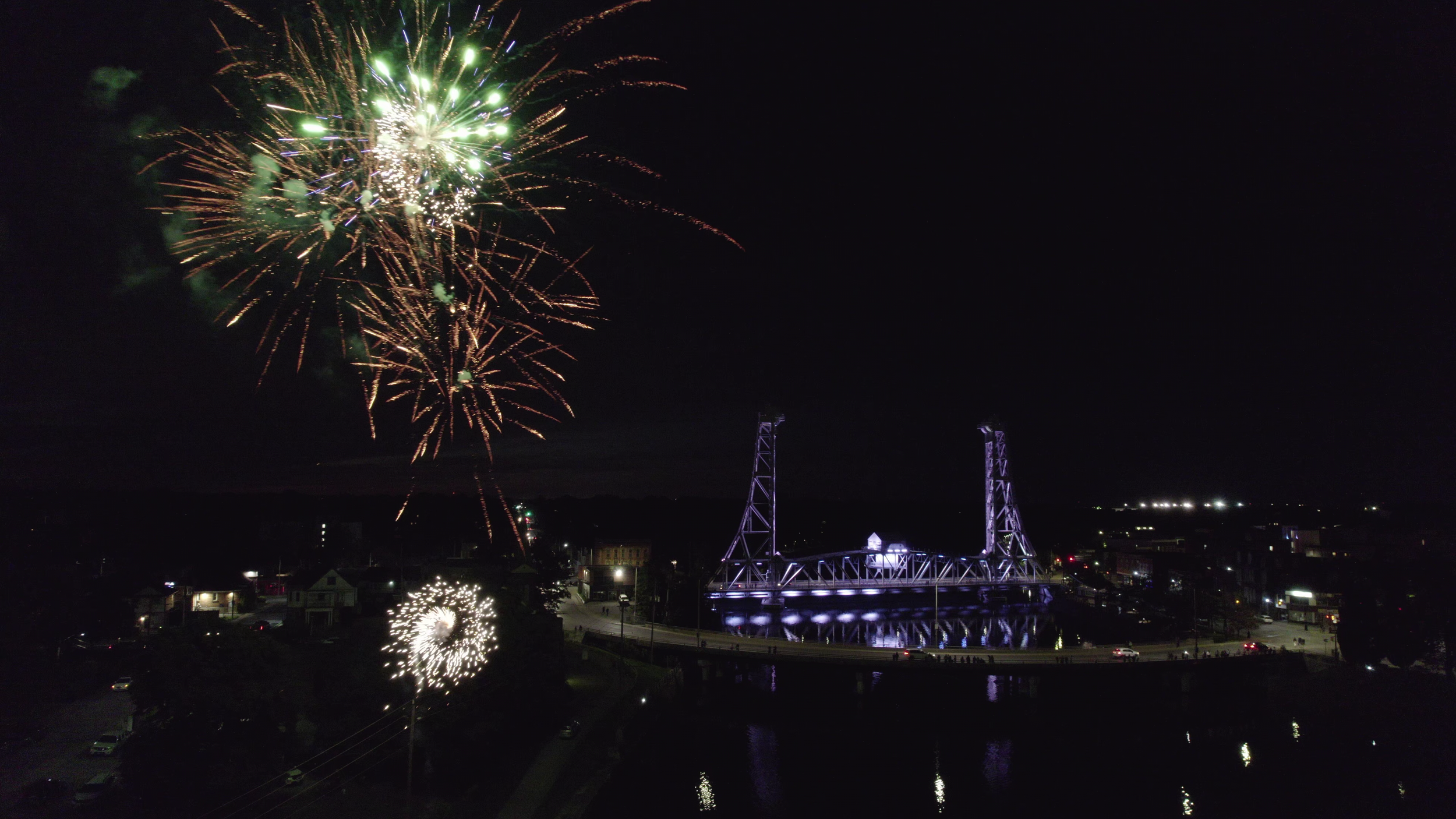 Nighttime fireworks display over a bridge illuminated with purple lights, with reflected lights on the water below and a dark sky background.
