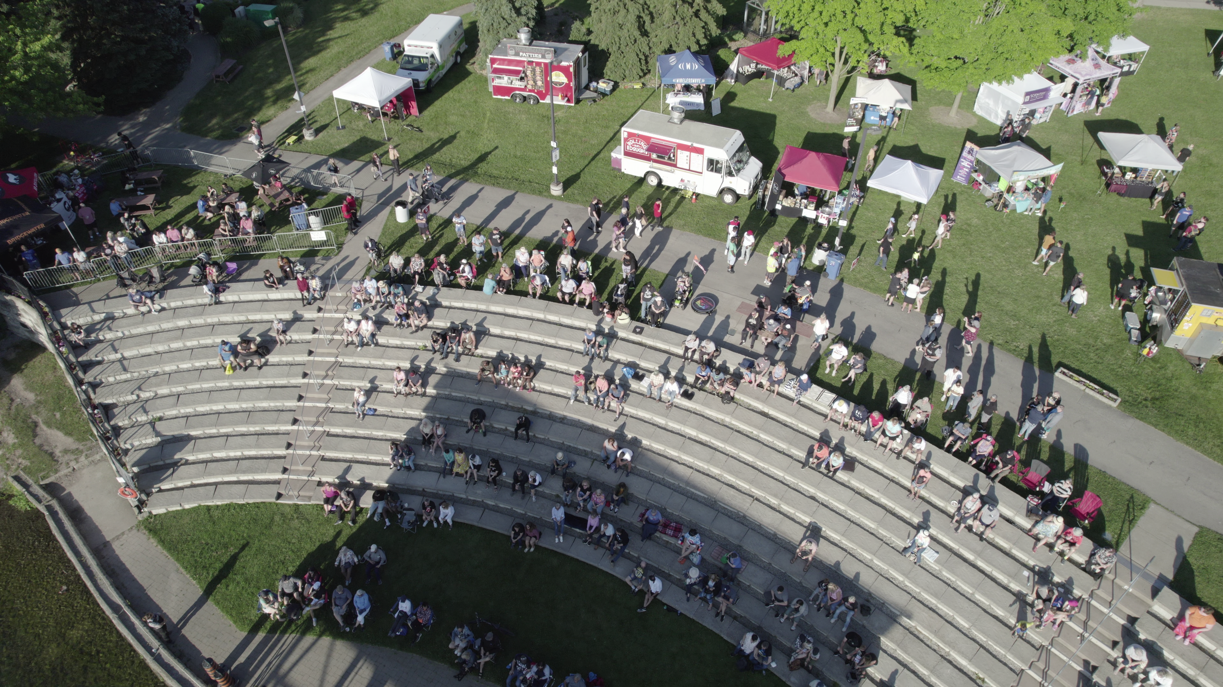 An outdoor amphitheater with about 50 people seated on curved rows of concrete seats, facing a stage, with some standing audience members. Behind the amphitheater, there is a grassy area with food trucks, tents, and people walking around under trees.
