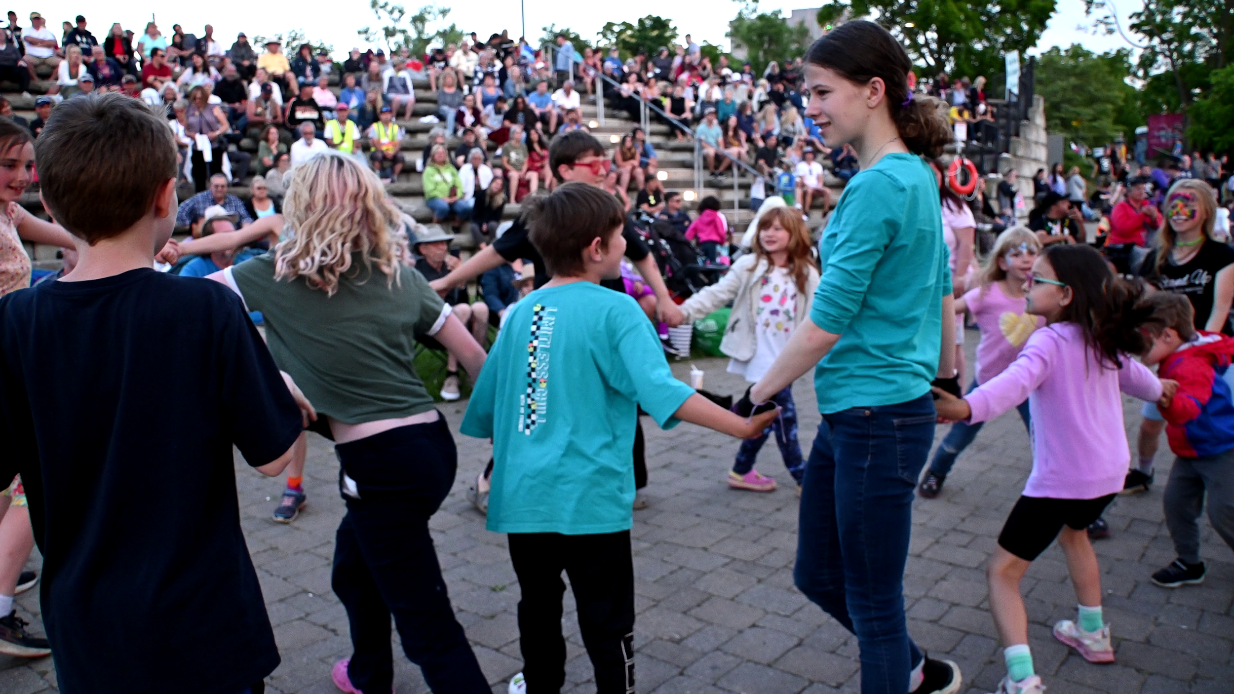 Children and adults dancing and playing in a circle outdoors at a public event, with a large seated crowd in the background.