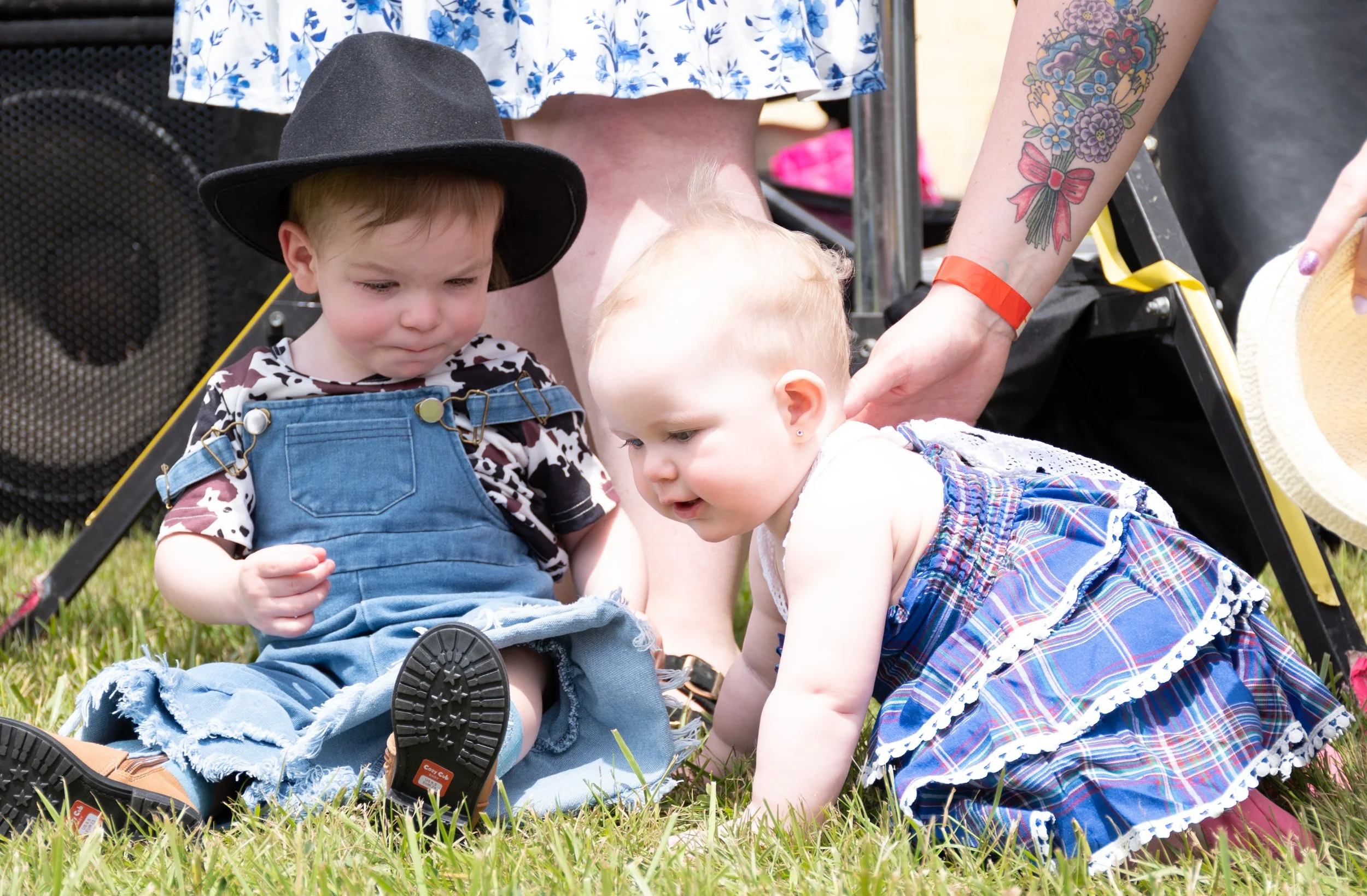 Two young children, one boy and one girl, sitting on grass during a sunny outdoor event. The boy wears a black hat and overalls with a leopard-print shirt, while the girl wears a plaid skirt and white top. Two adults are partially visible, one with t