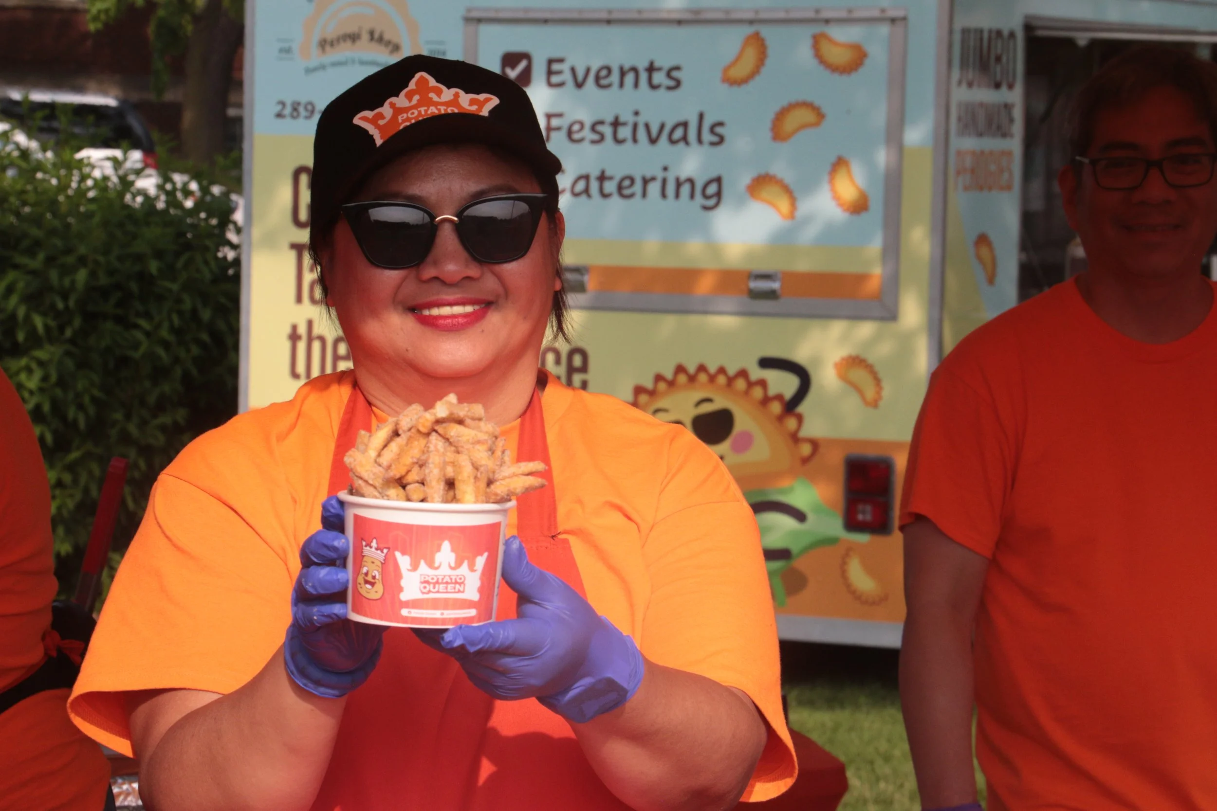 Woman wearing sunglasses, blue gloves, and orange shirt smiling while holding a cup of deep-fried potato strips at an outdoor food event.
