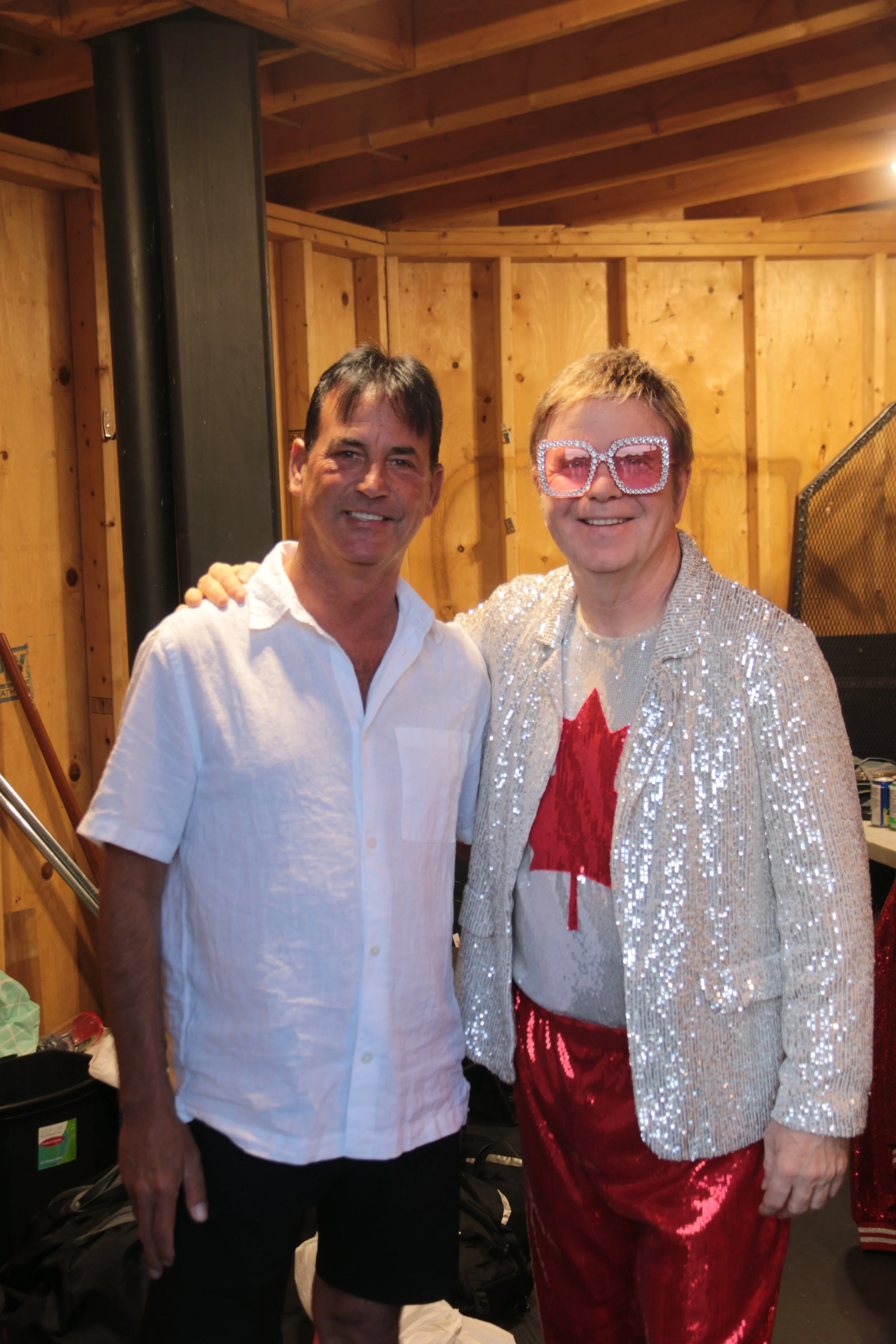 Two men standing together in a wooden room, smiling at the camera. The man on the right is wearing large sparkly glasses, a silver sequined jacket, a shirt with a Canadian flag, and shiny red pants.