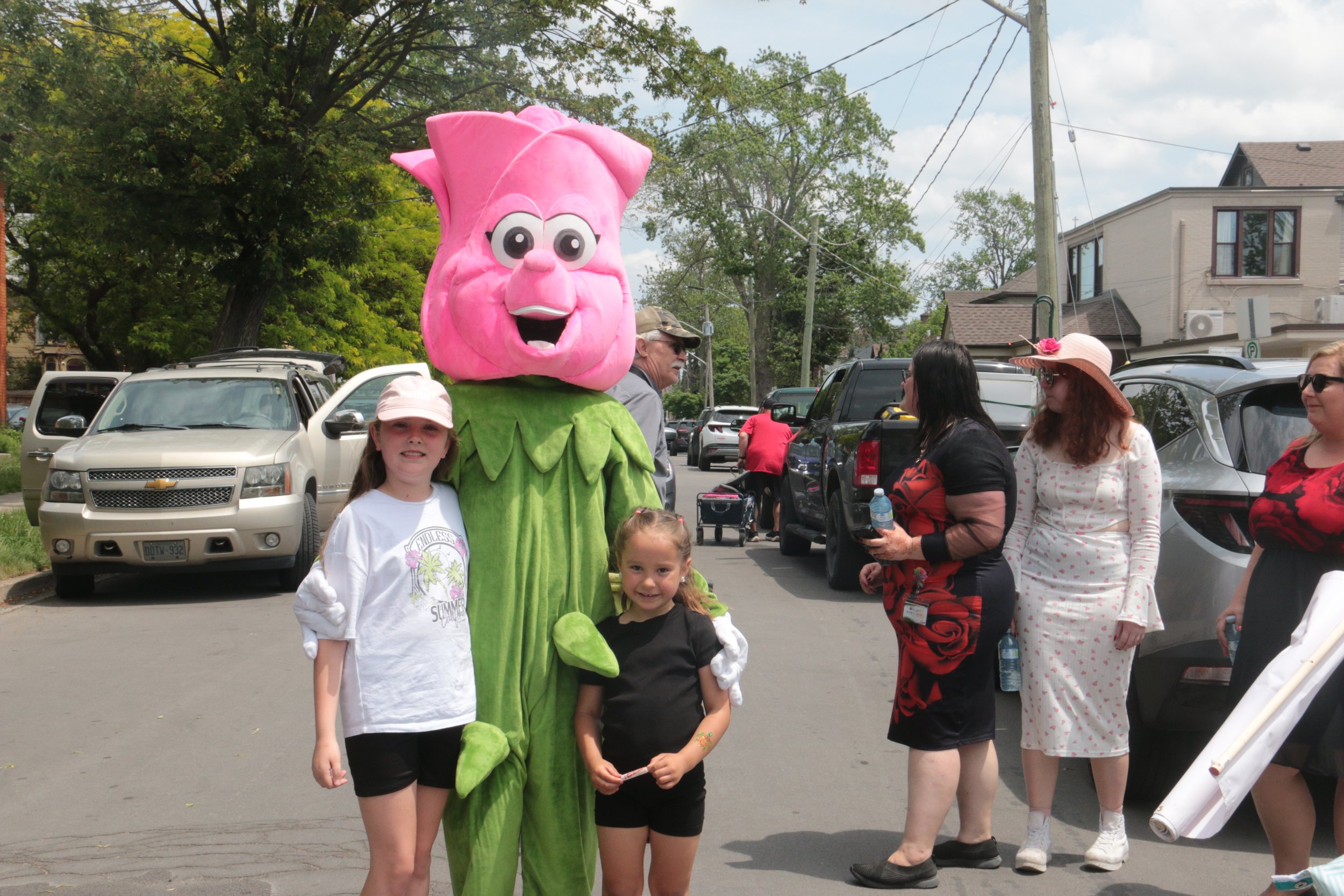 Children and adults smiling and taking pictures with a person in a pink anthropomorphic flower costume on a street with parked cars and houses in the background.