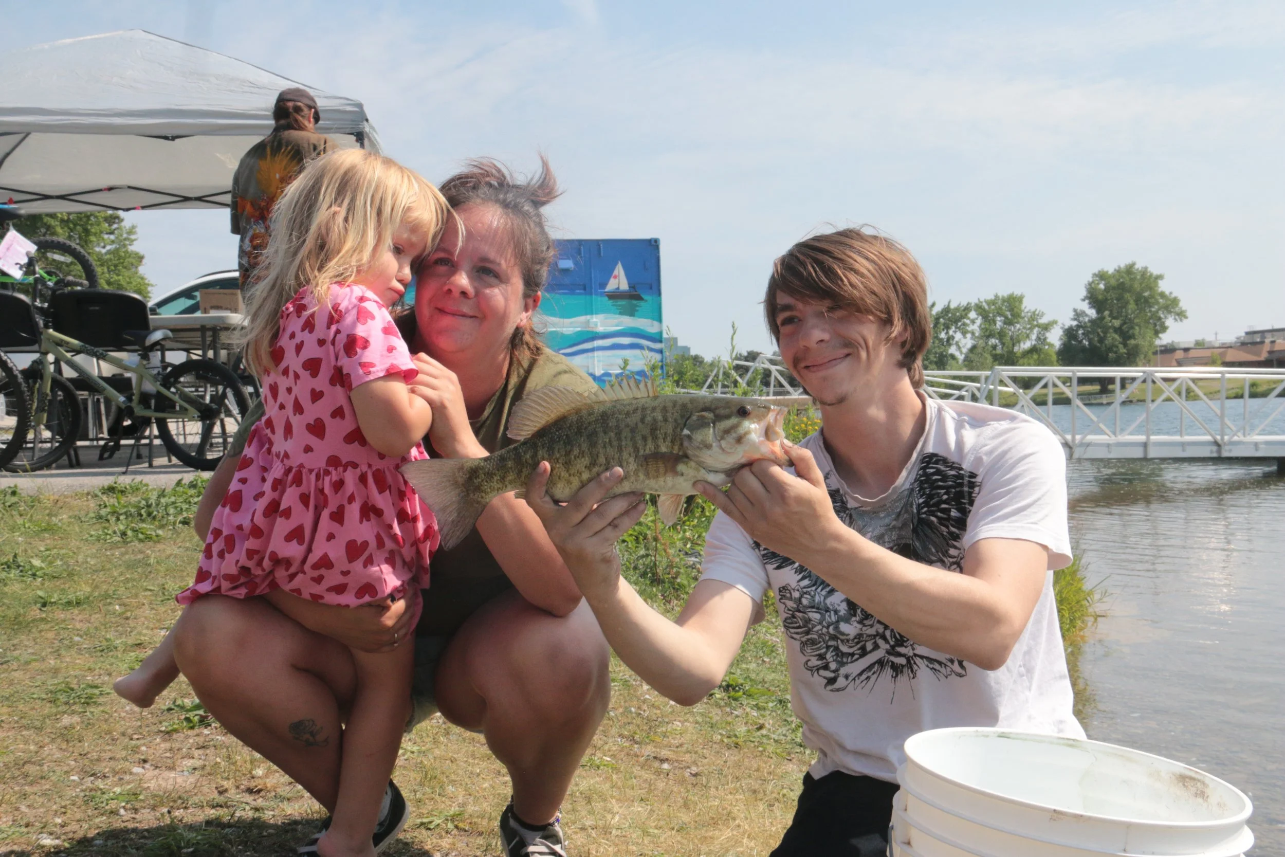 A family by a river, holding a large fish, with a young girl in a pink dress and adults smiling and posing.