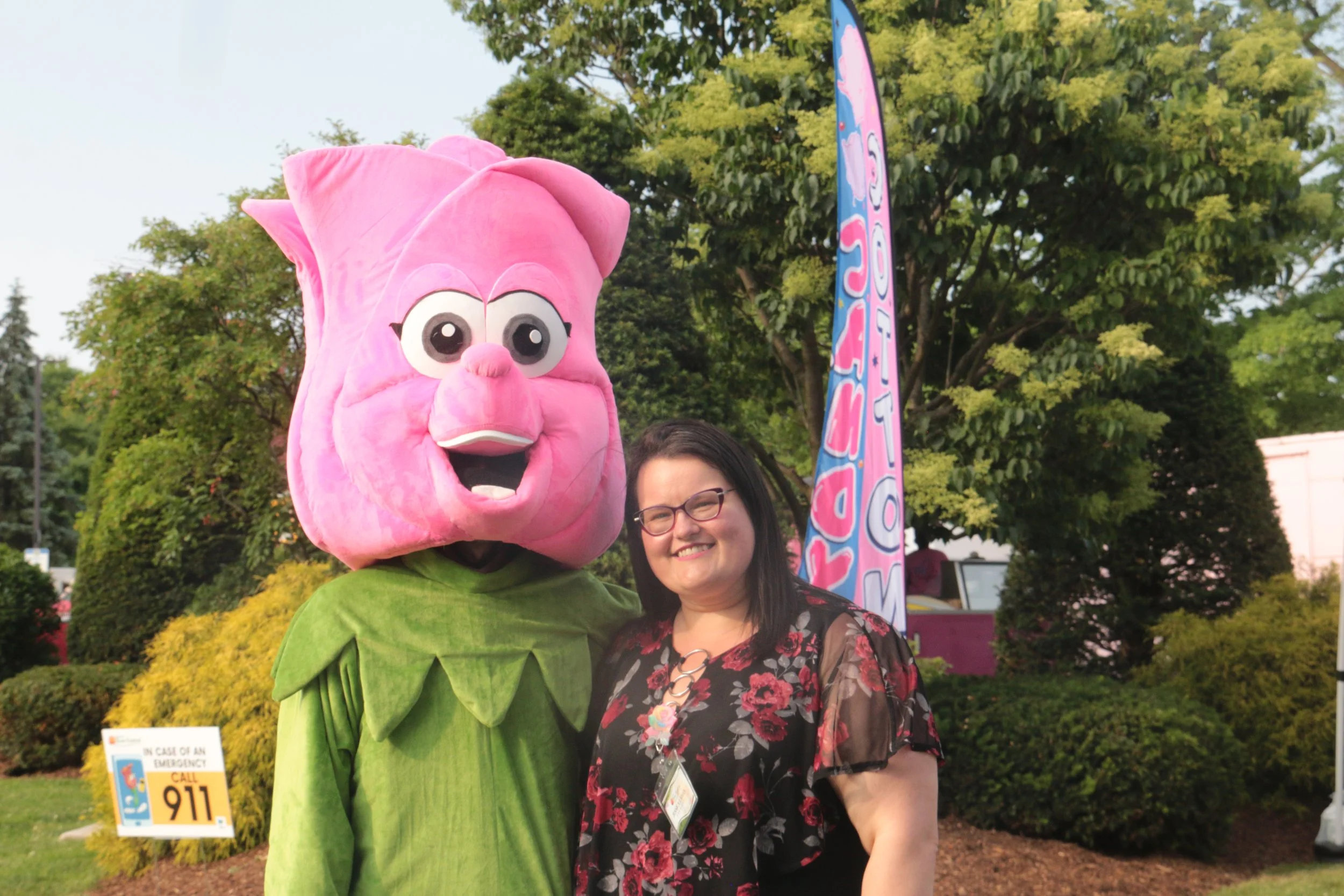 A woman standing next to a large pink pig mascot with big eyes, a snout, and a friendly expression, outdoors in a park-like setting with trees and colorful banners.