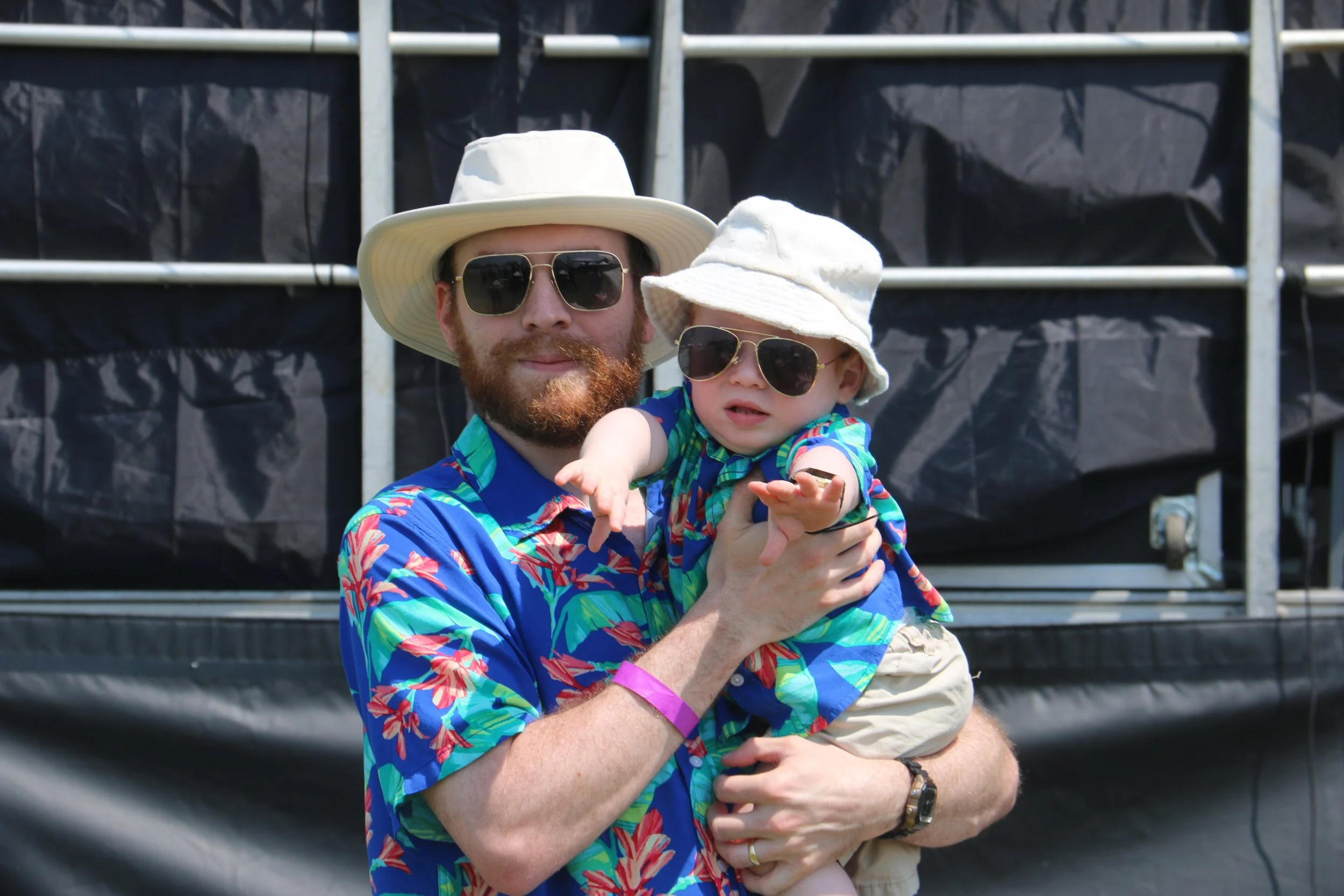 A man and a young boy wearing sunglasses and straw hats, dressed in tropical shirts, outdoors in bright sunlight.