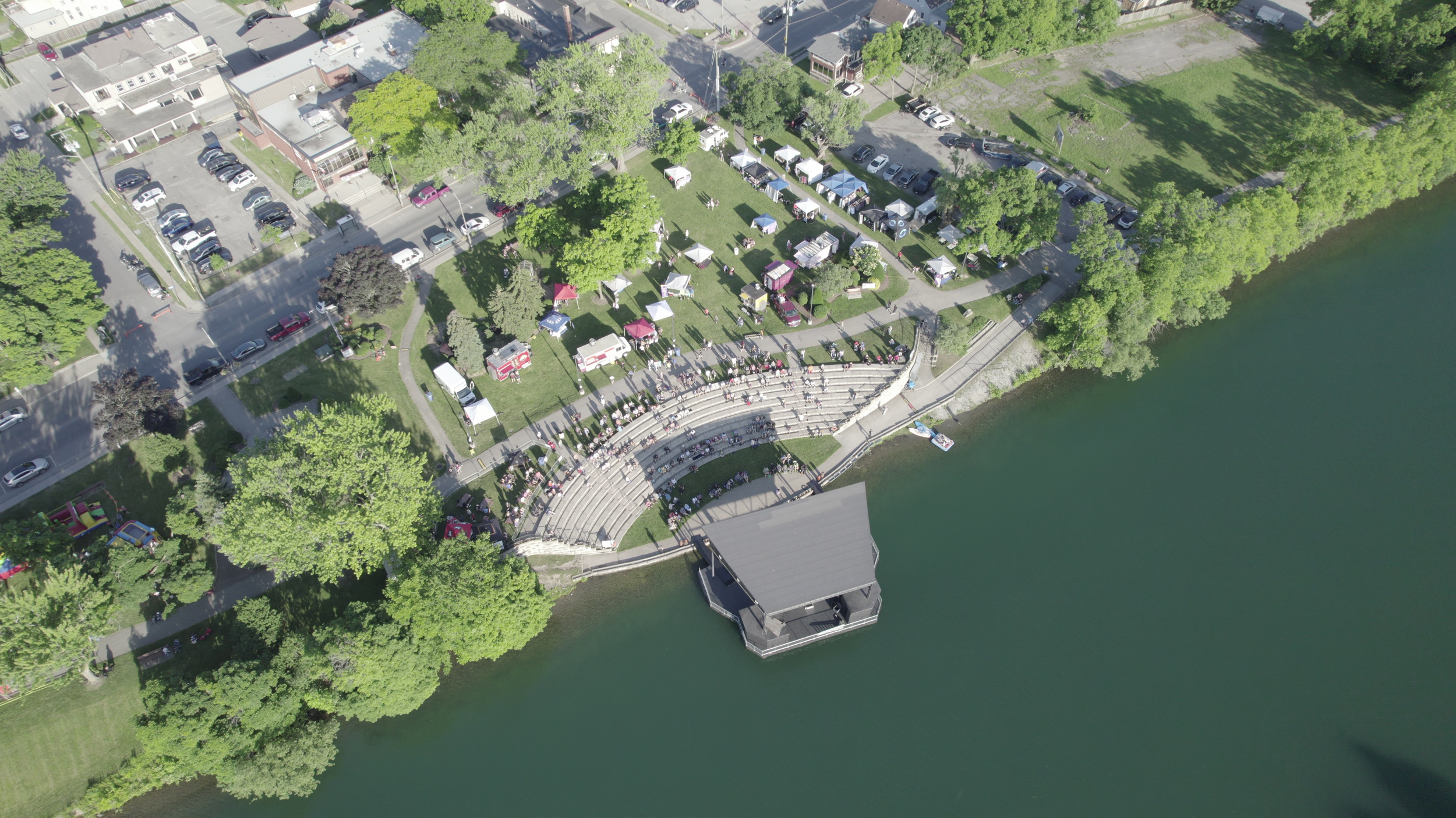 Aerial view of a lakeside park with a concert stage, outdoor seating, tents, and gathering people, surrounded by green trees and residential buildings.