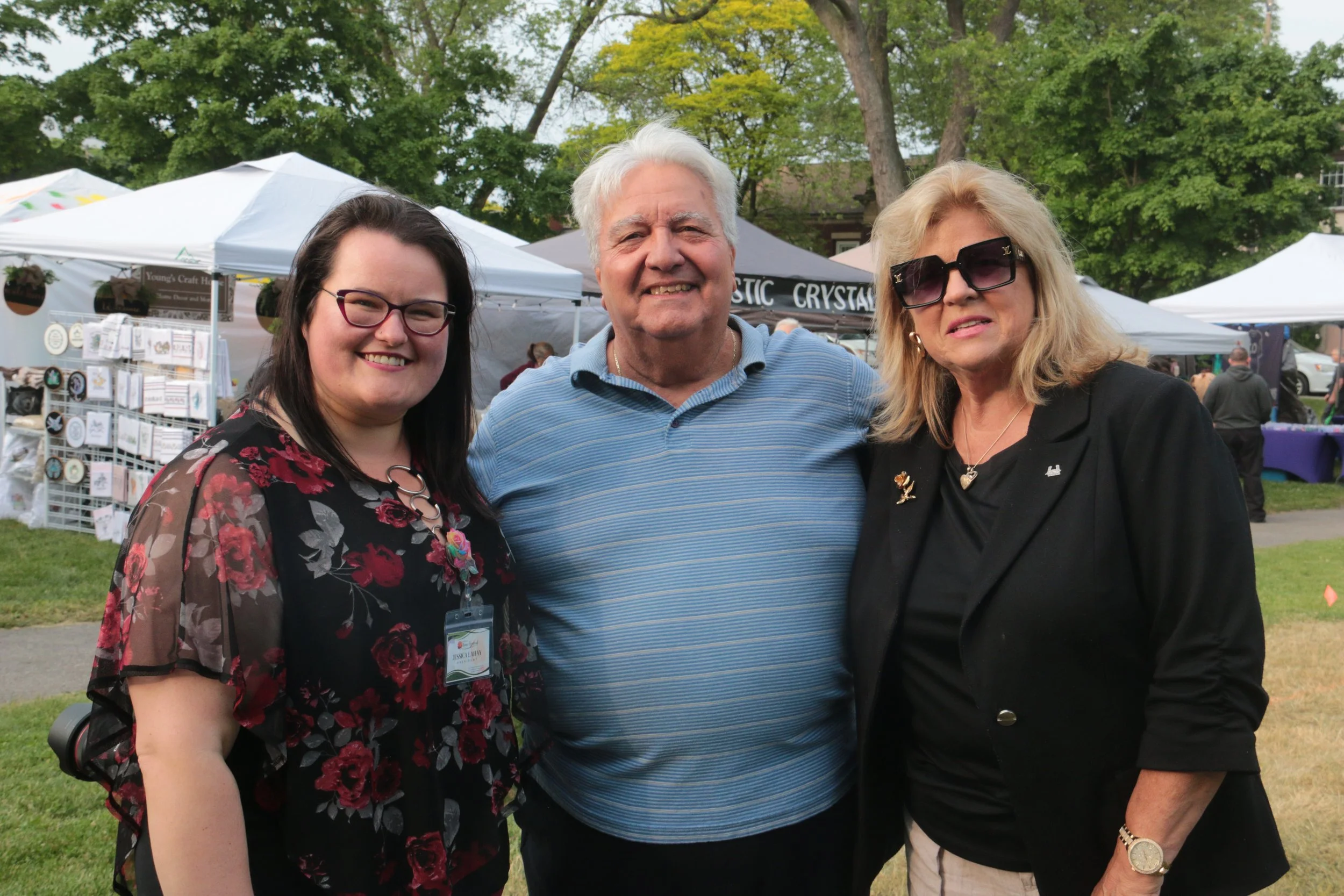 Three people standing together at an outdoor event with vendor tents and green trees in the background. The group includes a smiling woman with dark hair, glasses, and a floral top; a smiling older man with white hair in a blue shirt; and a woman wit