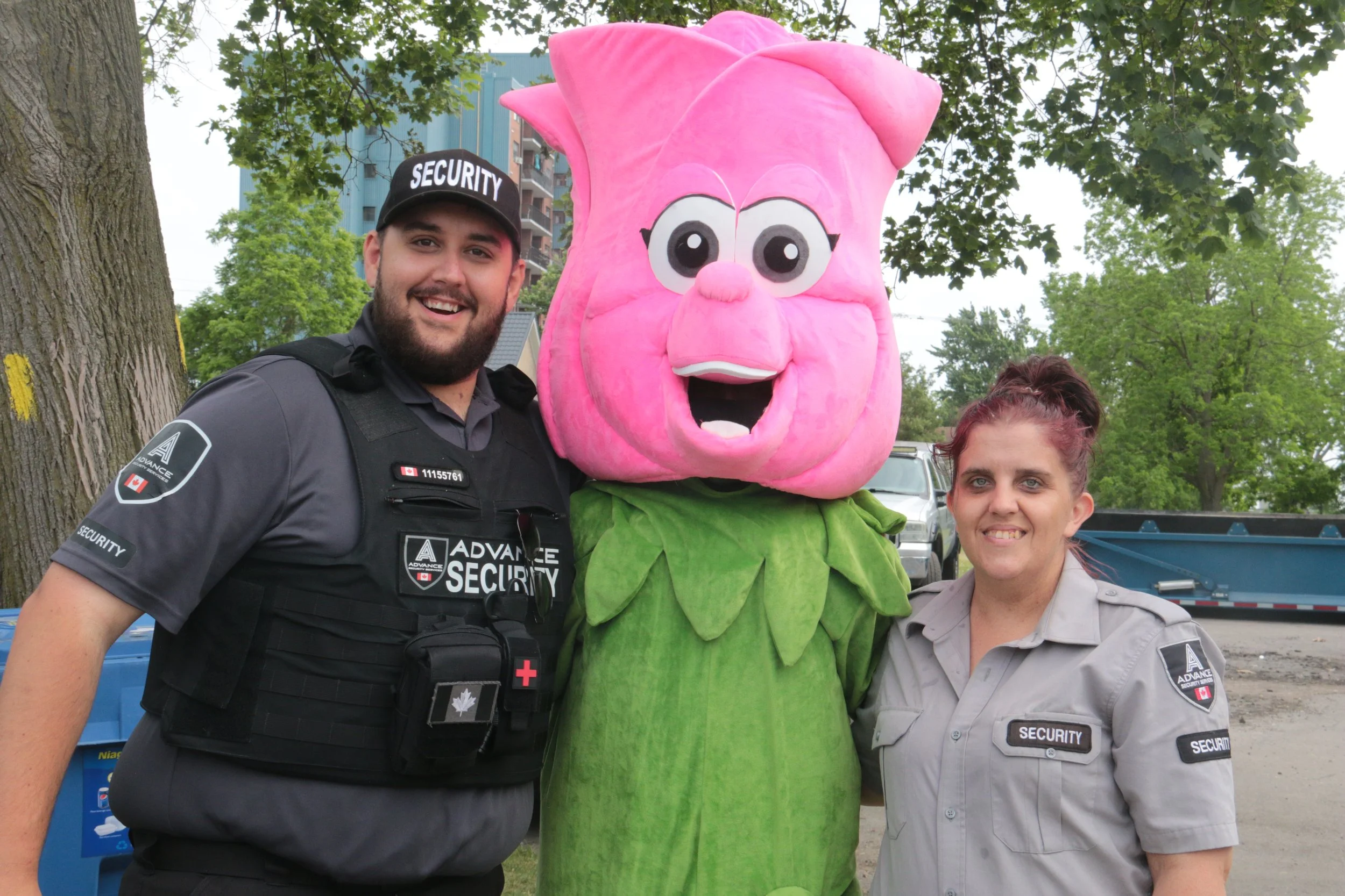 Two security personnel, a man and a woman, are smiling and standing next to a person in a large pink flower costume with cartoon eyes and a wide open mouth. They are outdoors with trees and parked cars in the background.