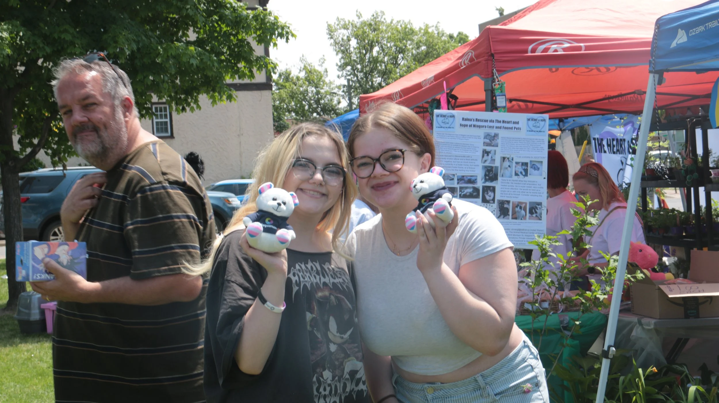 Two young women smiling and holding panda plush toys at an outdoor event, with colorful booths and other people in the background.