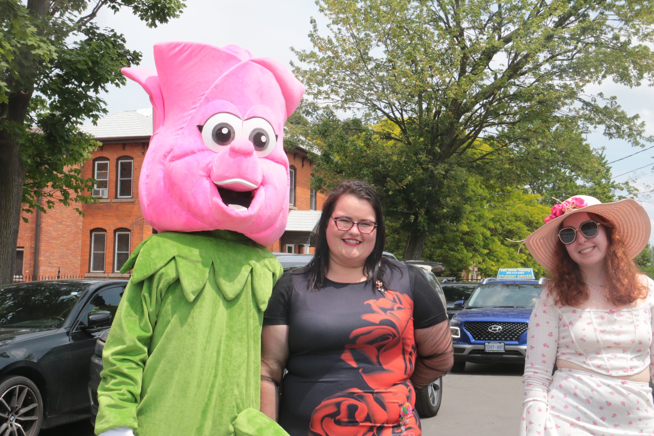 Three women standing outdoors, one in a pink flower costume, two smiling at the camera, with trees and parked cars in the background.