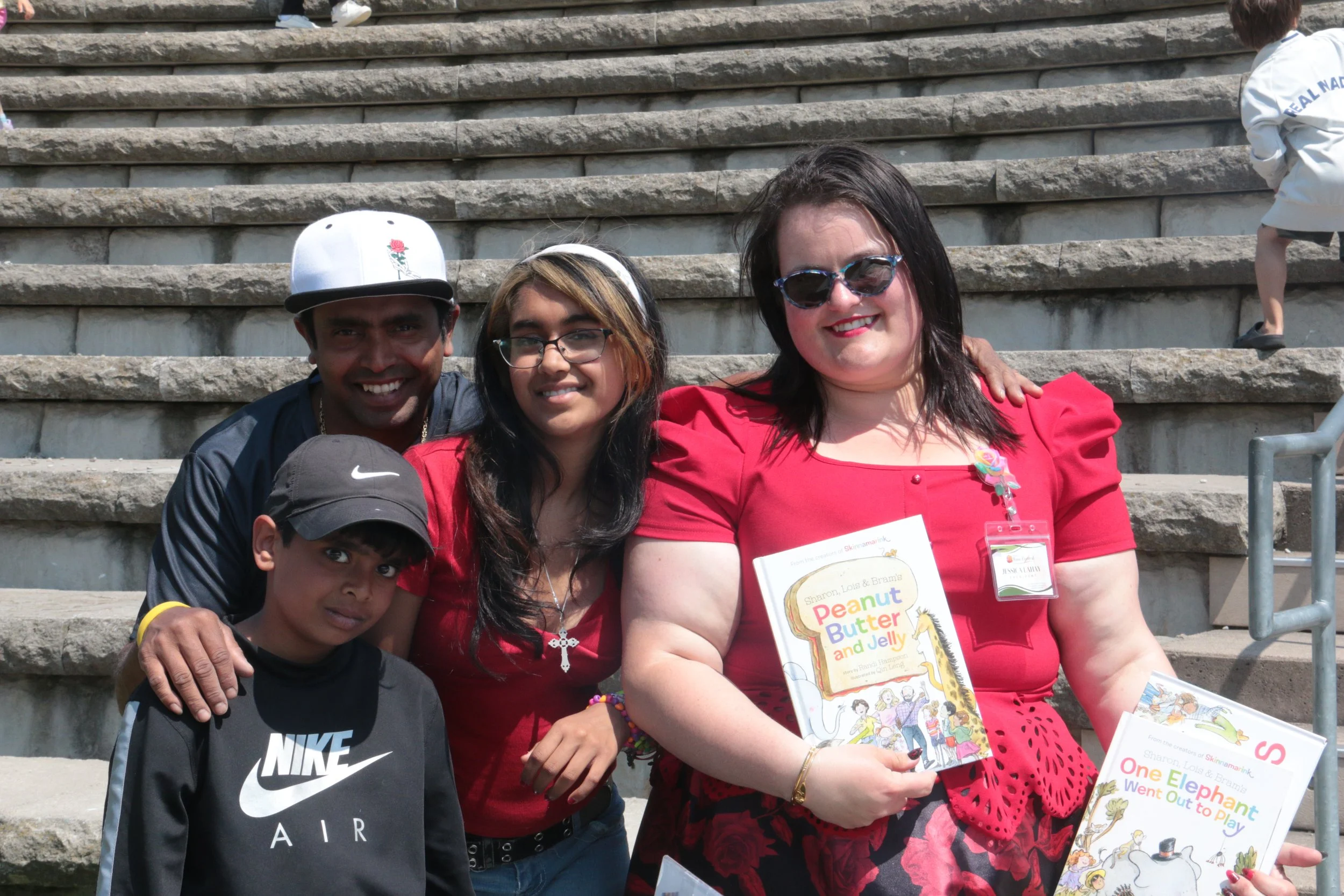 Group of five people, including children and adults, posing on outdoor concrete steps. The woman on the right is holding children's books, and the group is smiling at the camera.
