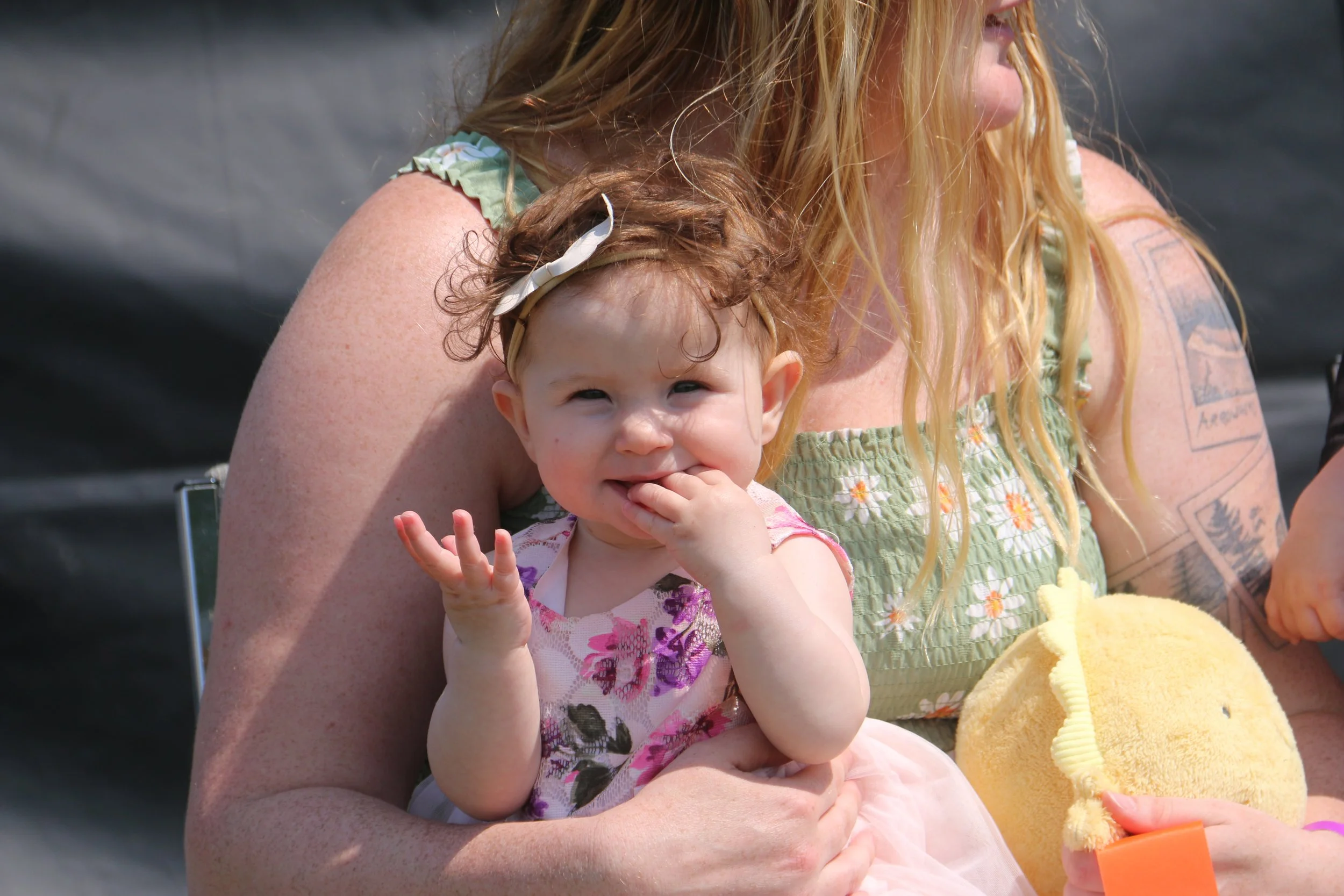 A smiling toddler girl with red hair, wearing a floral dress with a bow in her hair, sitting on a woman's lap outdoors.